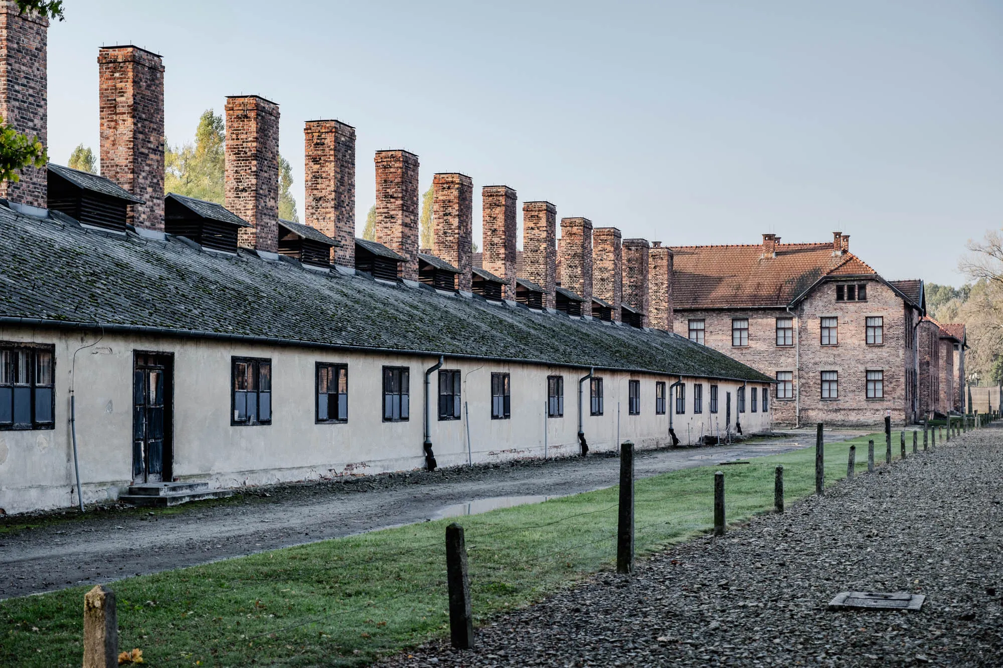 The image shows a long, low building with a row of tall brick chimneys on the roof. The building is made of concrete and has a slightly faded, pale yellow exterior. There are small, rectangular windows evenly spaced along the side of the building. In the foreground, the ground is a mixture of gravel and short, green grass. A row of wooden fence posts extends from the building out into the gravel, disappearing in the distance. There is a single, weathered wooden door in the middle of the side of the building, with a concrete stoop leading up to it. There is another building further in the distance. It is larger and taller, made of brown brick with a red tile roof. The windows are similar to the shorter building, except there are more of them. The top of the building is hidden behind a small forest of trees in the distance. The sky is pale blue and clear. The image is most likely taken from a side angle. The buildings appear to be old and weathered and have an ominous feel about them.  This photo depicts the Crematorium at Auschwitz-Birkenau, a Nazi concentration camp during World War II. 
