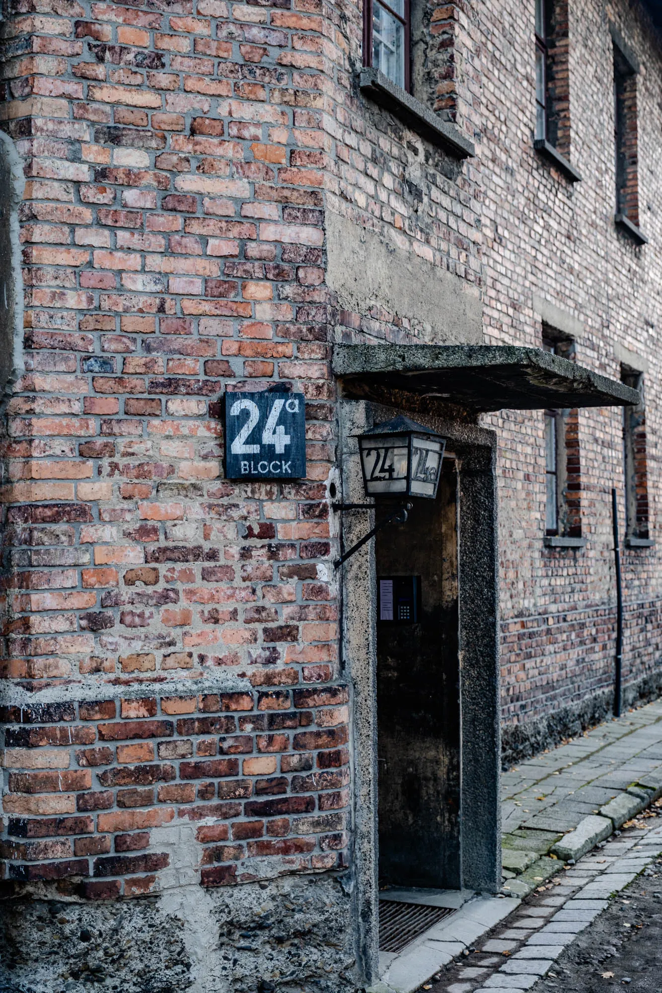 The image is of a brick building with a doorway. The building is made of red brick and has a few windows. There is a sign above the doorway that says "24a Block". There is a light fixture above the doorway, and the doorway is open and appears to lead into a dark room. The building is old and worn. There are some leaves and pebbles on the ground in front of the building.  This is a building at the Auschwitz-Birkenau concentration camp.