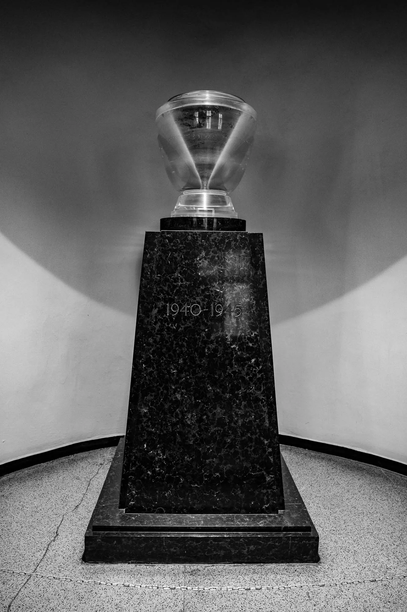 A black and white image of a black marble, square-shaped pedestal, with a large, clear glass, cylindrical, bowl-like structure on top.  The bowl-like structure has a  very subtle, ridged texture.  There are faint shadows cast from the structure. The pedestal is inscribed with the years "1940-1945".  The pedestal is set against a plain, light-colored wall. There is a thin, black, curved molding along the lower portion of the wall. The pedestal sits on a textured, gray, floor.  