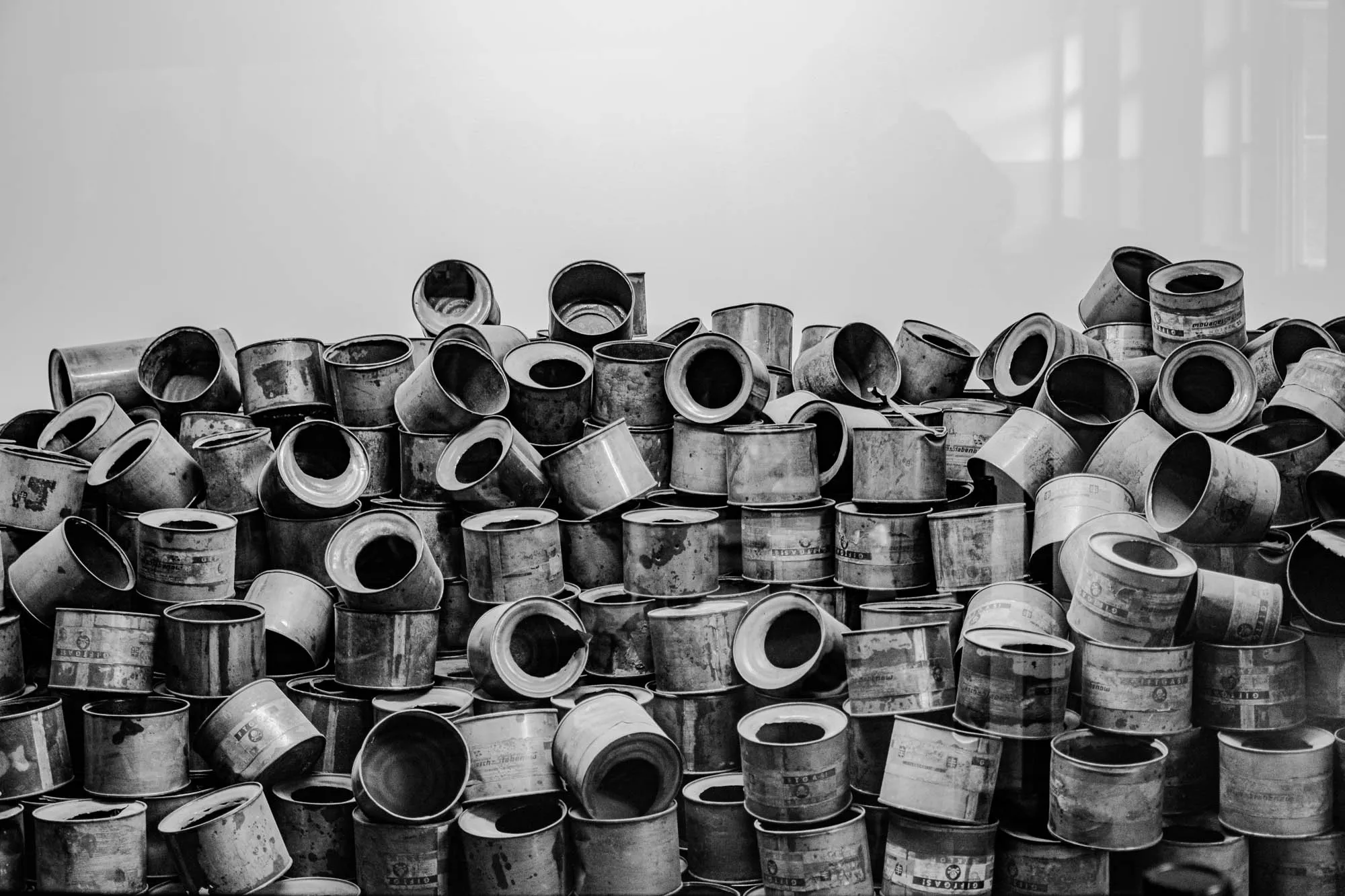 The image is a black and white photo of a large pile of metal cans. The cans are all different sizes and shapes, and they are stacked on top of each other in a haphazard way. Some of the cans have labels on them, but most are just plain metal. The cans are all covered in dirt and grime, and they look very old. There is a plain white background behind the pile of cans.  The photo is a stark reminder of the waste and destruction that can be caused by human activity.