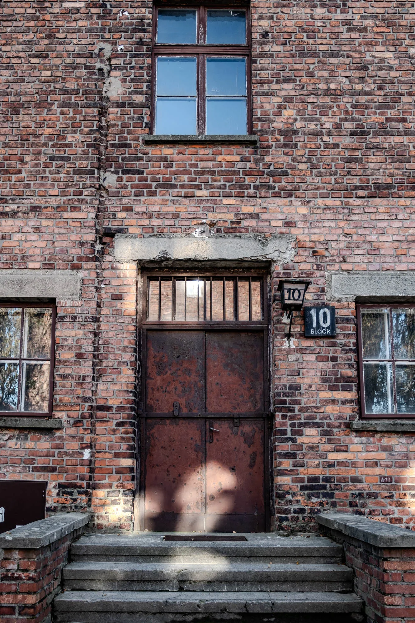 This image is of a brick building with a large, rusted metal door in the center. There is a window on either side of the door, and another window above the door. The building is old and worn, with signs of age and weathering. The door is closed, and there is a sign above it that reads "Block 10." Below the door are a set of concrete steps leading up to the building. The door appears to be secured with a metal latch. The window panes are all rectangular. The wall is made of red bricks with some mortar in between them. The windows are likely wooden framed. There is a light fixture on the wall to the right of the door.