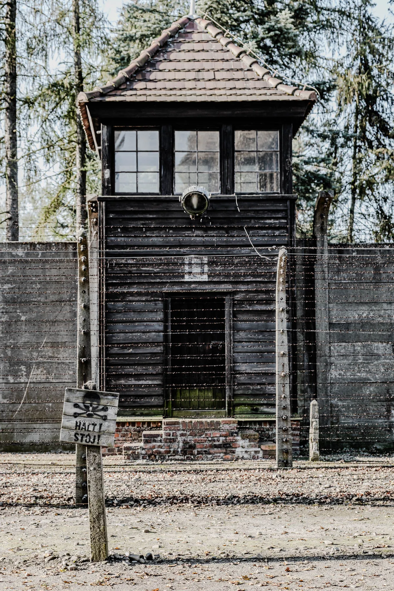 The image depicts a wooden guard tower with a red tile roof, situated within a barbed wire fence. The tower has two windows on the top level and a closed door below. The tower is made of dark wood, and a large light fixture is attached to its exterior. The barbed wire fence is made of multiple strands of wire, and the fence is attached to concrete posts. There is a sign in front of the fence that reads "Halt! Stoj!" with a skull and crossbones symbol. The ground is dirt, and there are fallen leaves scattered throughout the area. The photo appears to be taken on a cloudy day. The overall feeling of the image is one of confinement and despair, which is common imagery associated with concentration camps and places of oppression.  It's important to note that this image is a reminder of the atrocities committed during the Holocaust and should be viewed with respect and sensitivity. 
