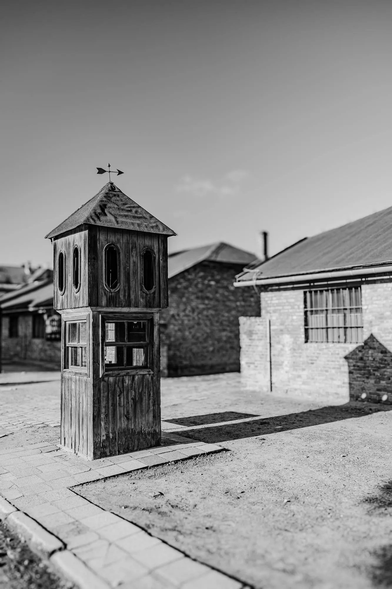 A black and white photo of a small wooden building with a roof and a weather vane on top, it has two square windows, it stands on a paved ground, behind the building is a brick building, with a small window, and a long shadow cast over the ground.