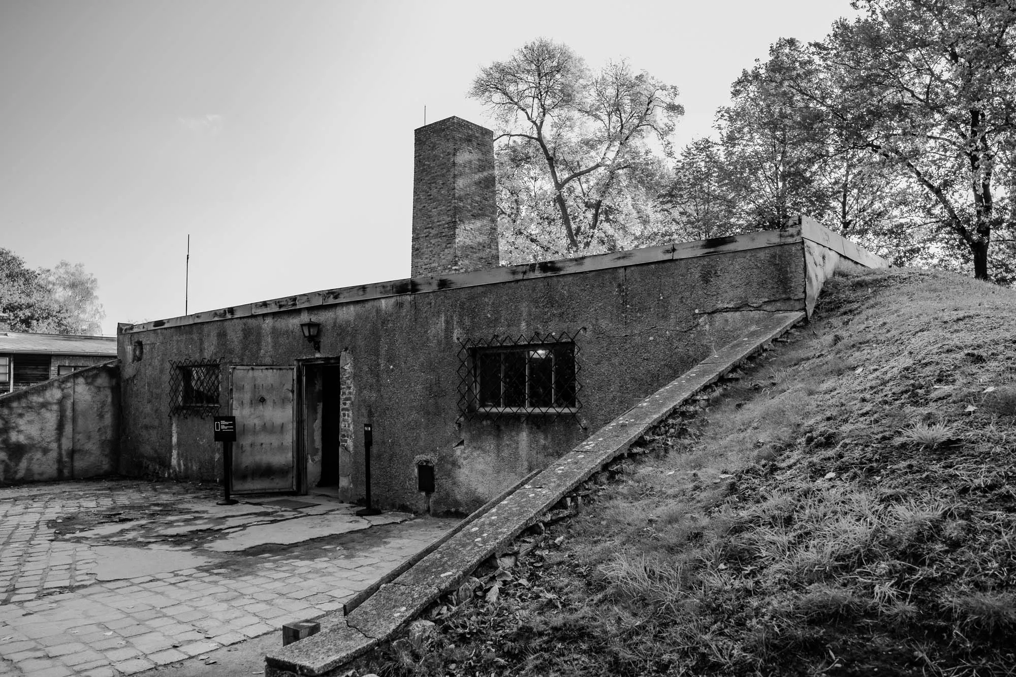 This is a black and white photo of a concrete building with a tall chimney.  The building has a large door in the front and a window to the right of the door. There are also some other windows along the side of the building. There is a concrete path leading up to the building, and a grassy hill in the background. The building is old and worn, and the grass is overgrown. The photo was taken on an overcast day.  This is an image of a crematorium in a concentration camp.