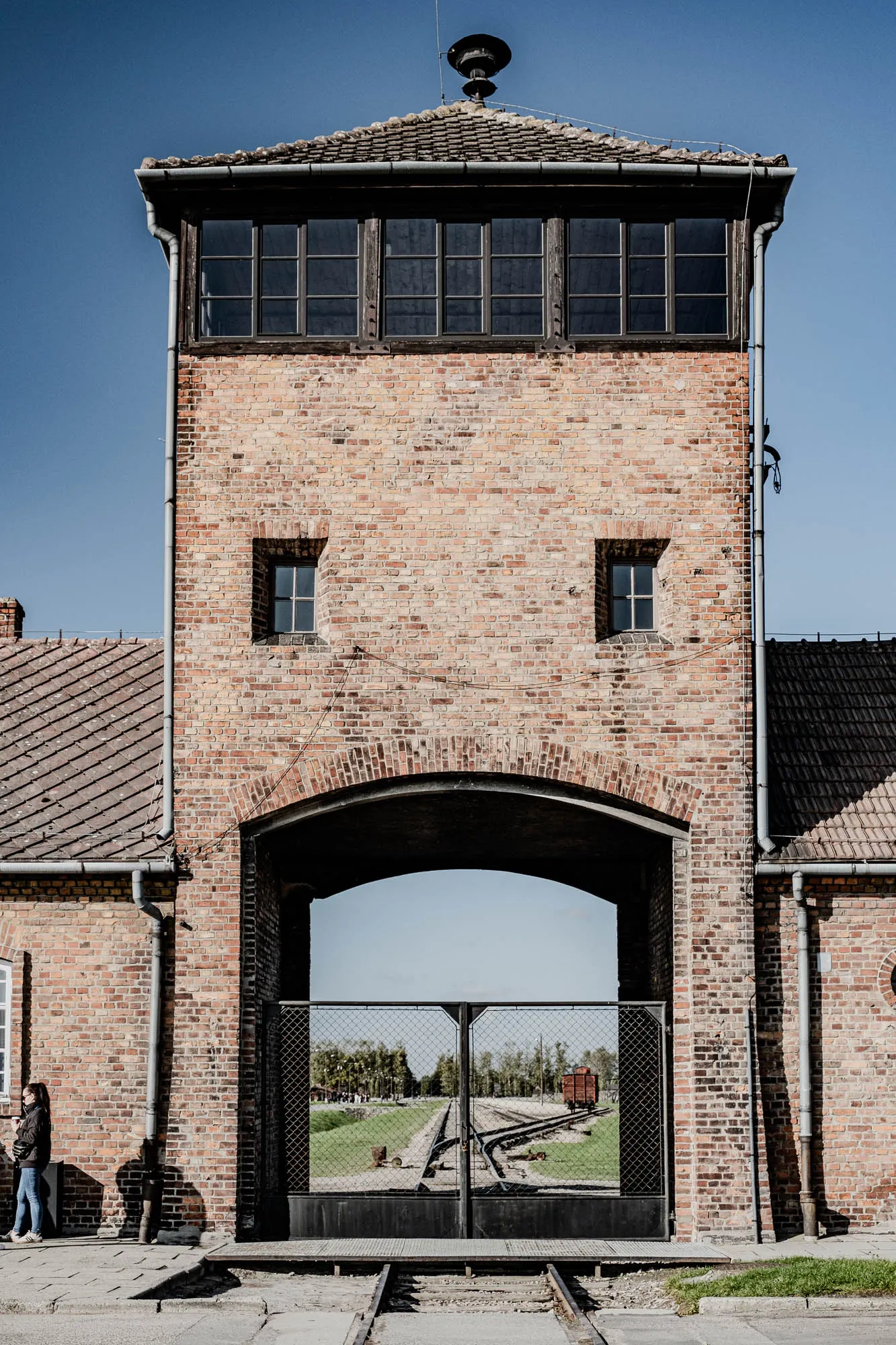 The image shows a brick building with a large arched gateway. The building is made of red brick and has a flat roof. There are two small windows in the building, one on each side of the gateway. The gateway is framed by a black metal archway. The gateway is closed with a black metal gate with a wire mesh. Behind the gate, you can see a grassy field with railroad tracks and a train car in the distance. There is a person standing in the foreground, to the left of the gate.  The building appears to be a part of a former concentration camp, with a somber and serious tone. 
