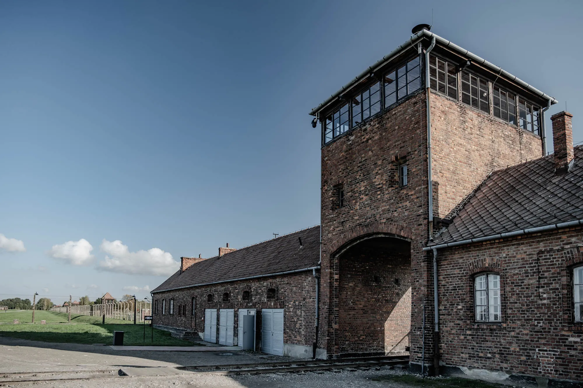 The image shows a brick building with a large arched doorway. The building is surrounded by a fence and there is a small building in the distance. The sky is blue and there are a few clouds in the sky. There is a long building with several windows and doors. It is a brick building with a slanted roof. The building has a large tower on the left side with a large window and a small window. The door is arched and made of brick. The building is set against a blue sky with a few clouds. The tower on the left is tall and has a large window on the top level and a small window on the bottom level. The brickwork is a little rough and the mortar joints are irregular. The tower has a metal roof with a gutter on the edge. The tower is built into the long building, forming part of the wall. The building has a long roof line and has a gutter on the edge. The windows in the long building are rectangular and are set into the brickwork. The windows are not very big. There are several doors in the long building. The doors are all rectangular and are made of wood. There are steps leading up to the doors. The ground in front of the building is made of dirt and gravel. There is a fence in the background and some trees. It is a somber scene, suggesting a place of imprisonment. The image is likely of a concentration camp.