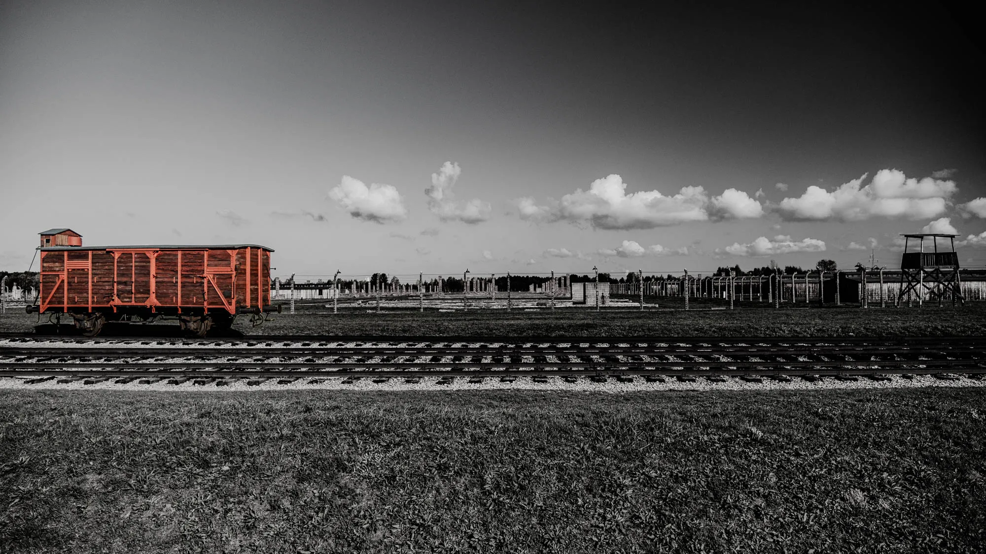 The image shows a single, red boxcar sitting on a set of train tracks in a field. The boxcar is facing the viewer, with the tracks extending into the distance. The rest of the image is in black and white, with the boxcar the only color in the picture. The tracks lead to a fence in the distance, with an empty guard tower in the upper right corner of the image. The sky is a gray color with white clouds.