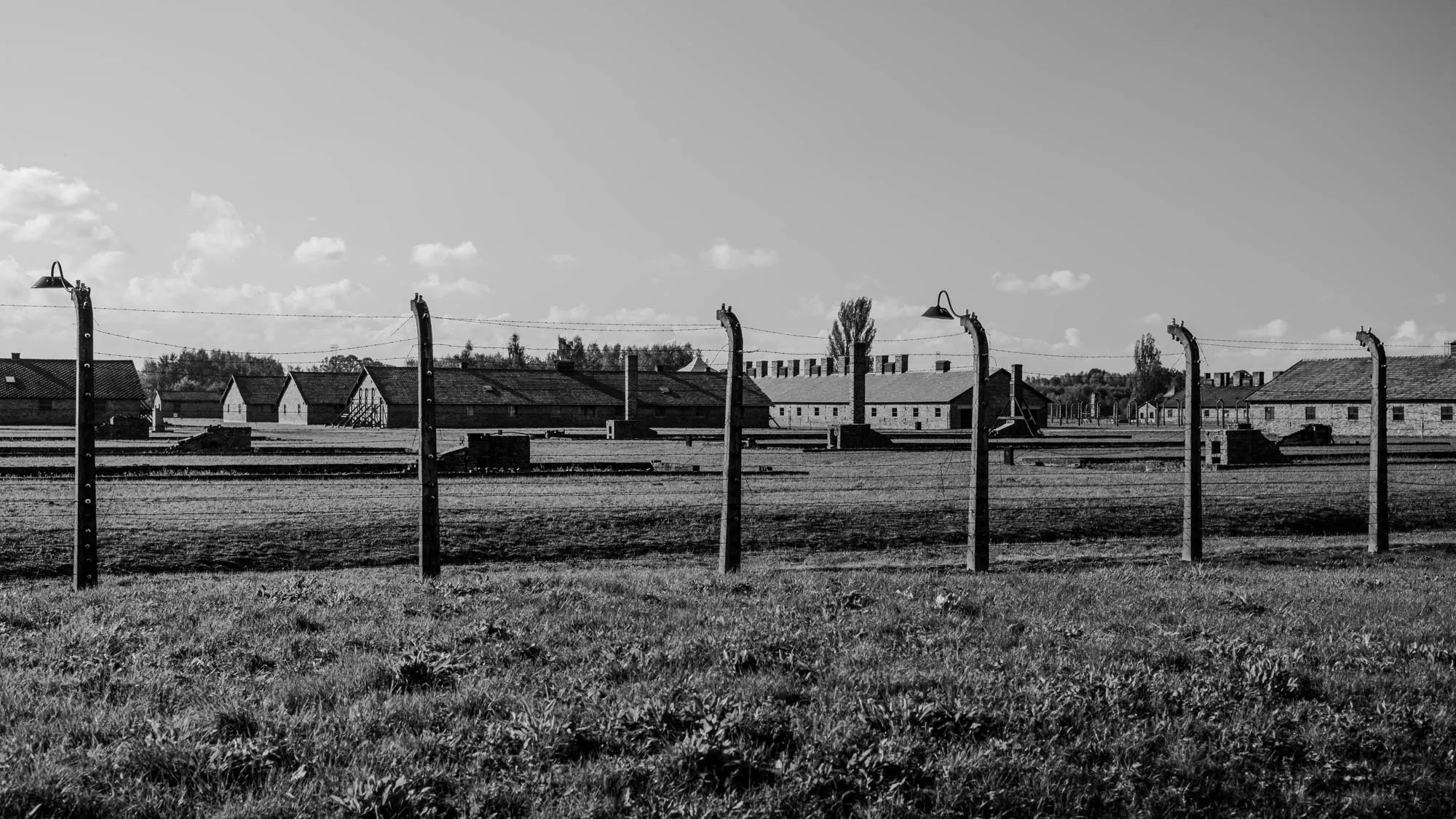 The image shows a black and white photograph of a concentration camp. The photo is taken from the outside, looking through a barbed wire fence. The fence is made of tall, wooden posts with several strands of barbed wire running between them. There are several tall, brick buildings behind the fence. There is a light fixture on top of each post in the foreground. The buildings are in the middle ground of the image and there is a short grassy field in the foreground. The sky is grey and cloudy. The image is a reminder of the horrors of the Holocaust and the importance of remembering those who were lost.