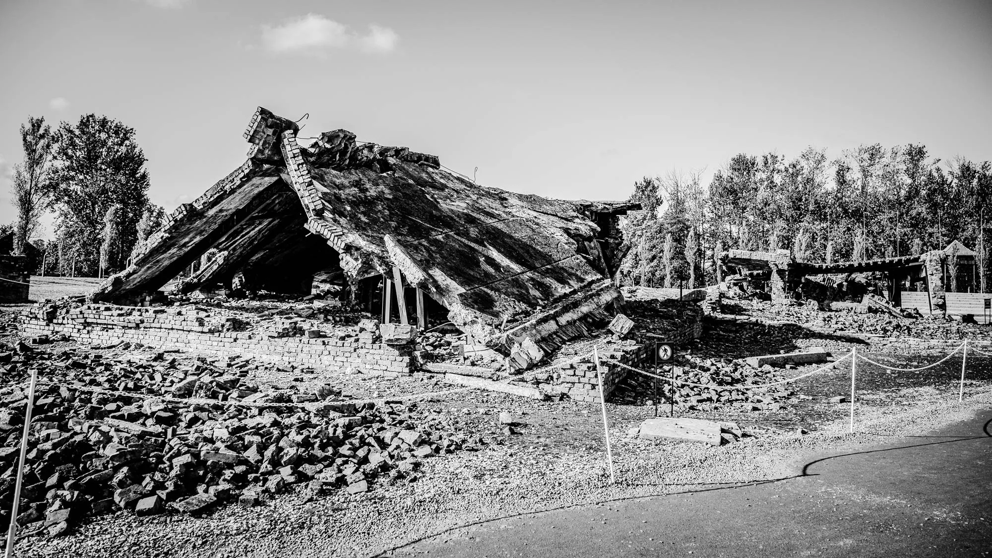 The image shows a black and white photo of the ruins of a building. It appears to be the remains of a brick structure with a collapsed concrete roof. The roof has fallen on the building, creating a large pile of rubble. The building sits in a field of broken bricks and stones. There are trees in the background, and a small, black-and-white sign with a symbol of a person walking can be seen on the right side of the image, near a white rope barrier that runs along the edges of the field.  The scene is very desolate and gives a sense of the destruction and loss that occurred.  The ruins appear to be a site of historical significance, possibly a war memorial or a place that was destroyed in a conflict.