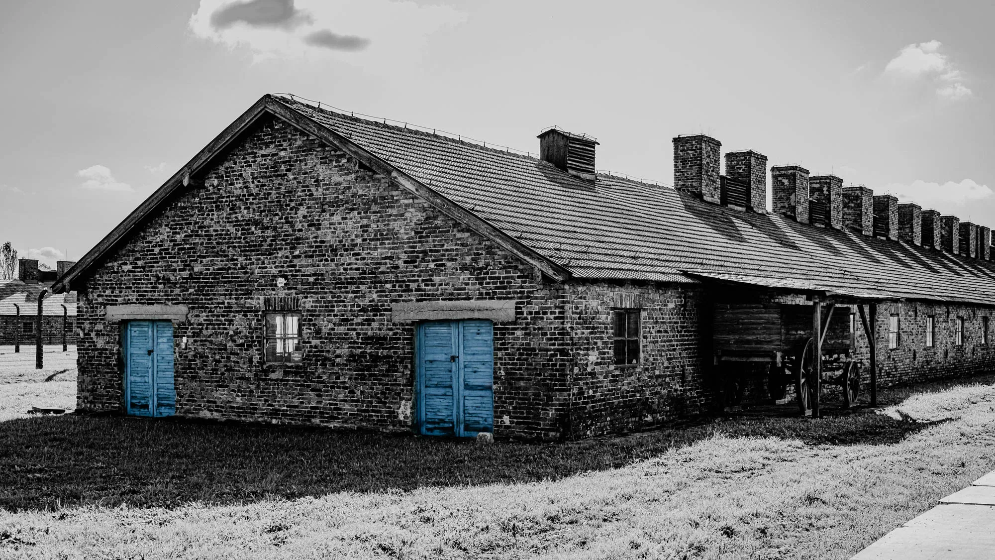 A black and white image of a long, brick building with a pitched roof. There are multiple chimneys and a small window on the side of the building. There are two wooden doors on the side of the building. The building is set in a grassy field with a paved path running in the foreground. There is a small wooden shed with a cart in front of the building. The sky is clear with a few clouds. This image is likely a photograph of a former concentration camp building. 
