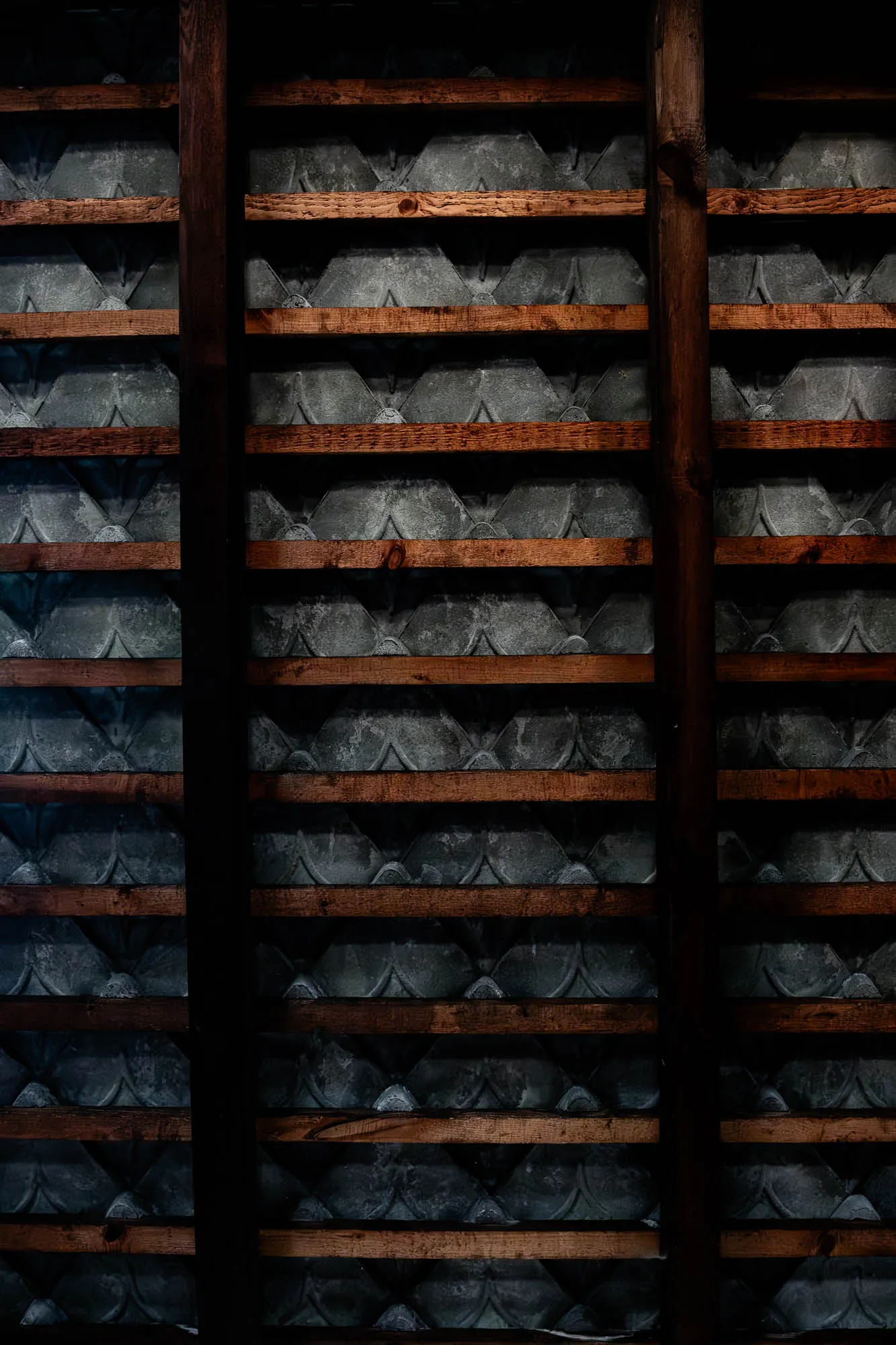 This is a close-up image of a wall with a unique design. The wall is made up of dark grey, slightly textured, tiles that are arranged in a repeating pattern of rounded triangles. The tiles are separated by horizontal wooden beams, which are slightly darker in color. There are two thicker wooden beams that run vertically, dividing the wall into three sections. The beams cast shadows on the wall, giving the image a sense of depth. The image is mostly dark, with only a few highlights on the wooden beams and the tiles.