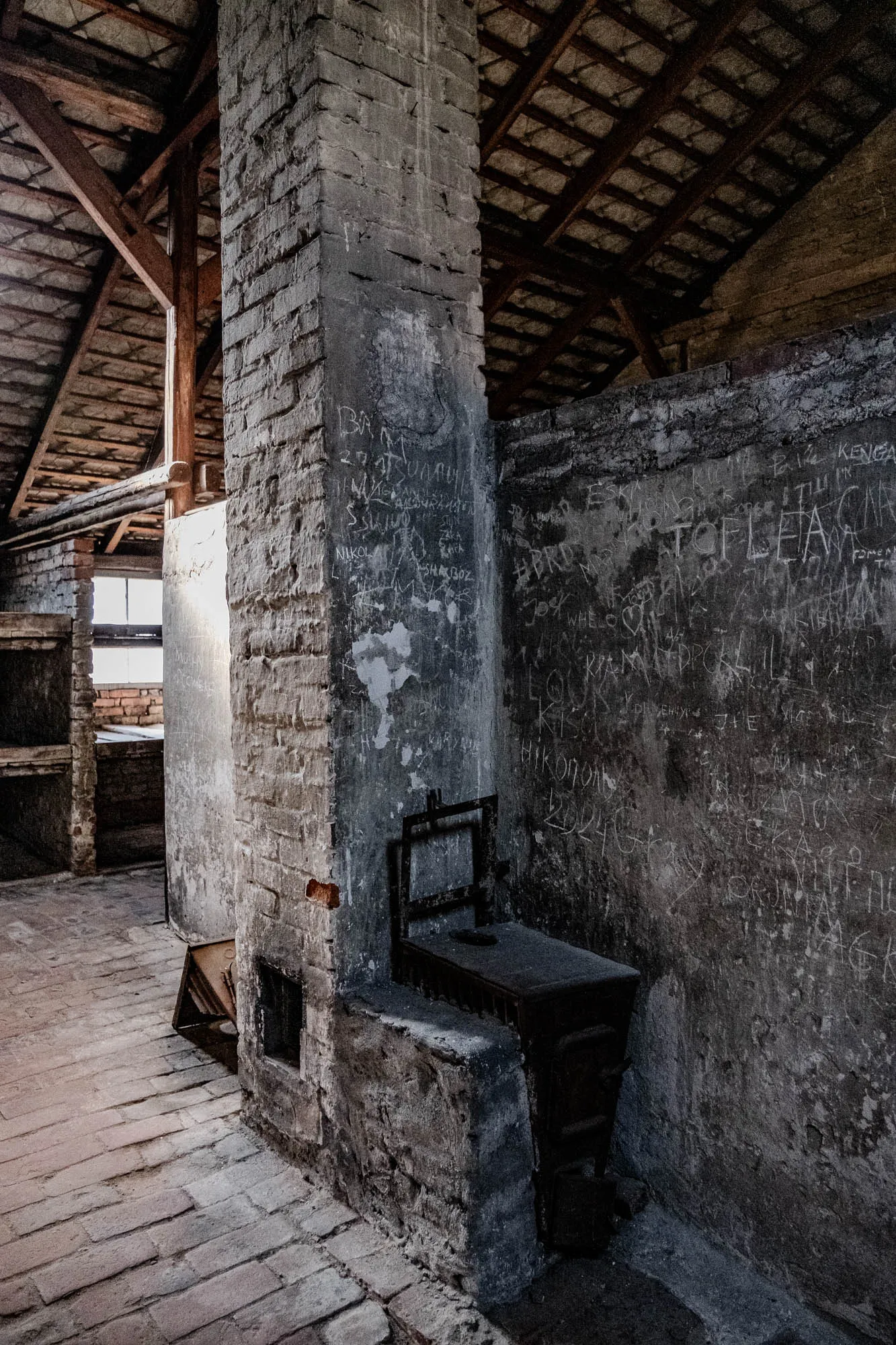 The image shows a dark, grungy room with an old brick chimney and a wood-burning stove in the corner. The room is lit by a single window that lets in a stream of sunlight. The walls are covered with writing in white chalk, including a variety of names and dates. The roof of the room is made of wood and has a grid pattern. In the center of the room, there is a brick path that leads towards the camera. There is a metal grate over the stove opening.