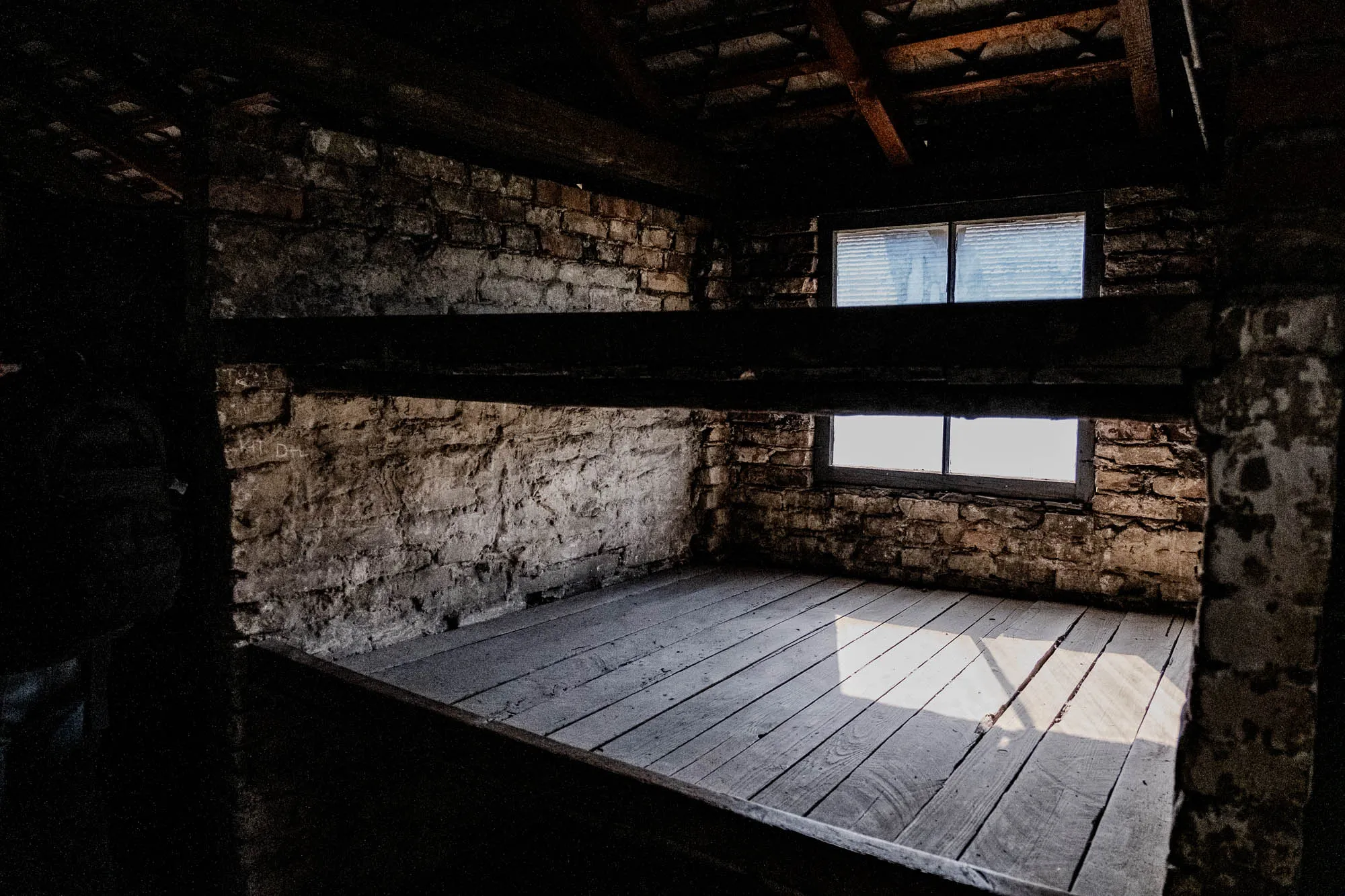 The image shows a dark, narrow room with a brick wall on one side. The floor is made of wooden planks, and there is a wooden bunk bed above the floor. The bunk bed is dark and appears to be empty. There is a window on the brick wall. The window is divided into two panes, and it is letting in a beam of light that falls across the floor. The room is otherwise dark and shadowy. The image may be of a prison cell or a similar type of austere, minimalist living space.