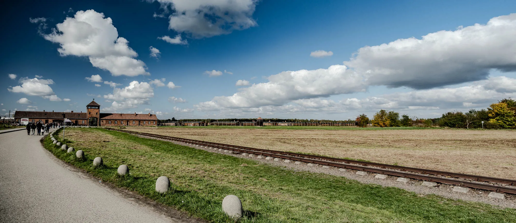 The image shows a wide shot of a grassy field with train tracks leading towards a brick building in the distance. The sky is blue and cloudy.  There are groups of people walking on a path to the right of the train tracks.  In the distance, there are trees lining the horizon.  The building has a watchtower in the center.   The image is taken from a low angle, looking up at the watchtower and sky.  The image is likely taken at a historical site. 
