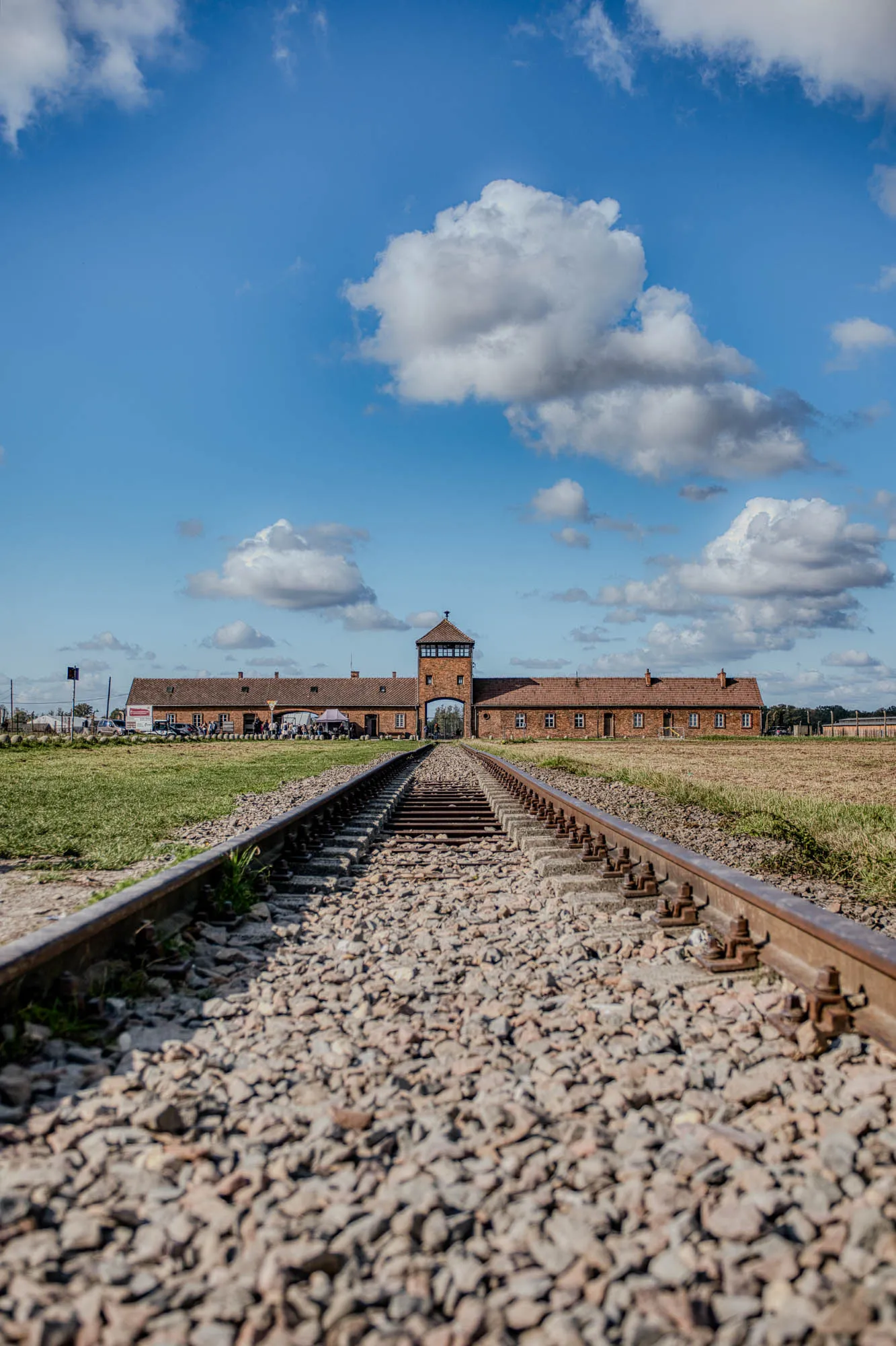 The image shows a set of train tracks receding into the distance. The tracks are made of rusty metal and are set in a bed of gravel. The tracks are surrounded by a field of green grass. In the distance, you can see a brick building with an arched doorway. The building is surrounded by a fence and there are people visible inside of it. The sky is blue and there are fluffy clouds. The image is likely taken from a low angle, making the tracks appear to converge in the distance.  This scene evokes a sense of history and somberness, as the train tracks were used to transport people to concentration camps during World War II. The image is a reminder of the horrors of war and the importance of remembering those who were lost.