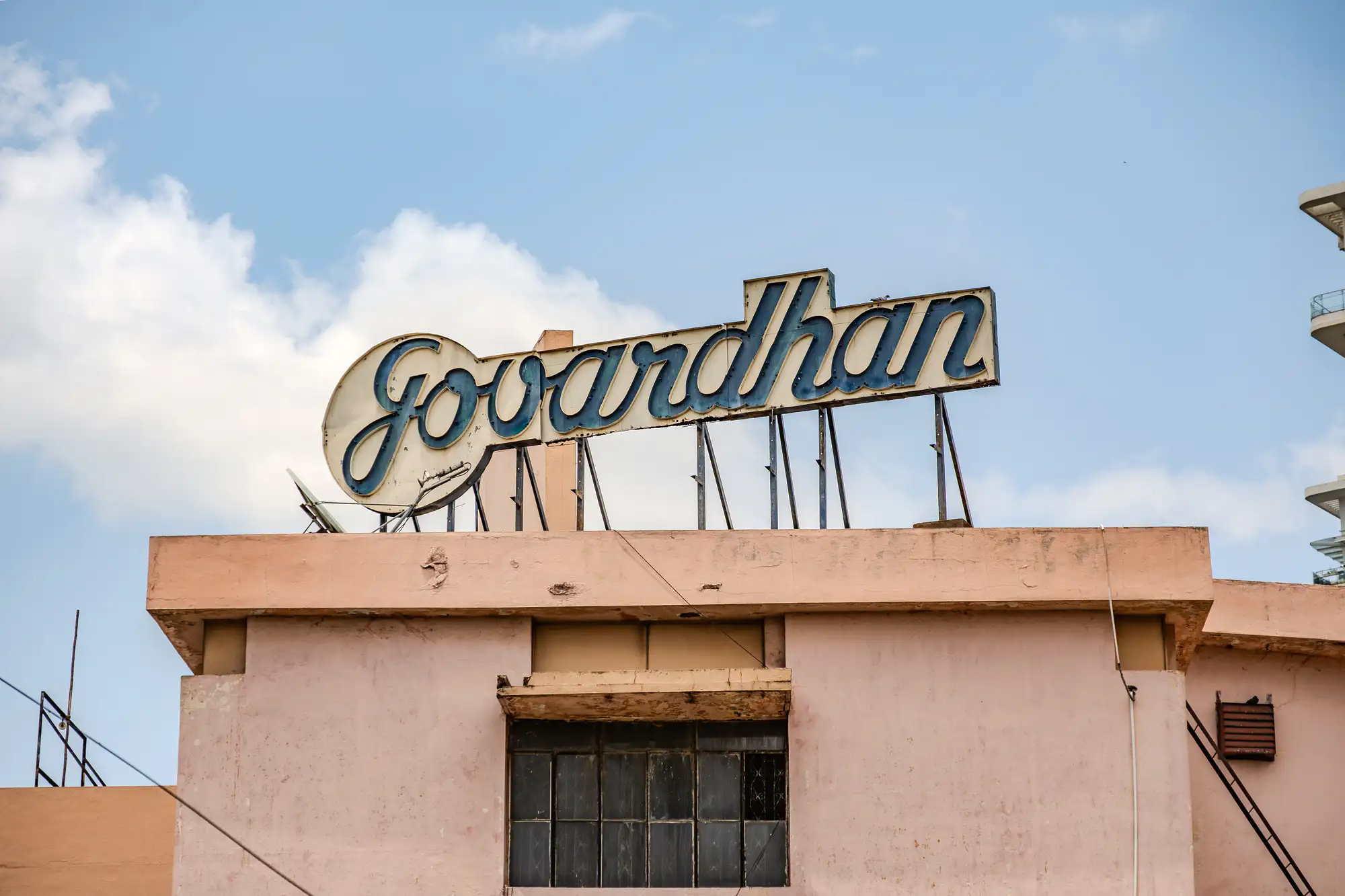A weathered vintage sign spelling out 'Govardhan' in blue cursive lettering sits atop the flat roof of a muted peach-colored building. Slender metal struts support the elevated sign against a daytime sky patterned with soft white clouds. Below the roofline, a multi-pane rectangular window is set into the worn stucco facade, accented by small architectural fixtures and structural wiring. The building's faded pinkish exterior contrasts subtly against the vibrant blue open air above.