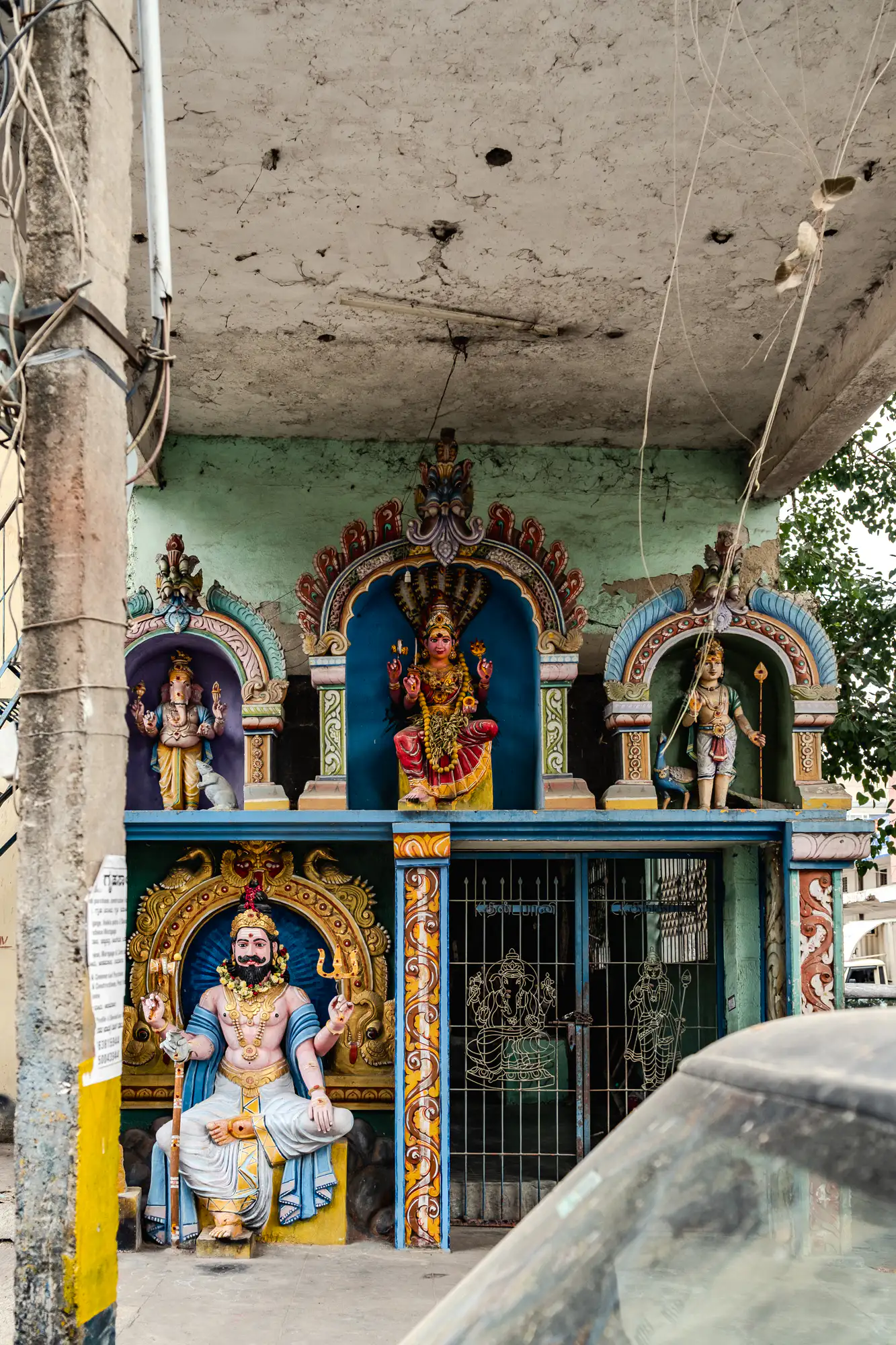 A multi-tiered street shrine stands integrated into the facade of a weathered urban building. Three colorful arched alcoves on the upper level display painted statues of Hindu deities, featuring Ganesha on the left, a multi-armed goddess beneath a serpent hood in the center, and Murugan with his peacock on the right. Below, a large mustachioed deity holding a trident sits majestically beside a gated entrance patterned with sacred silhouettes. A concrete utility pole laden with tangled wiring frames the left, while the edge of a parked car enters the foreground on the right.