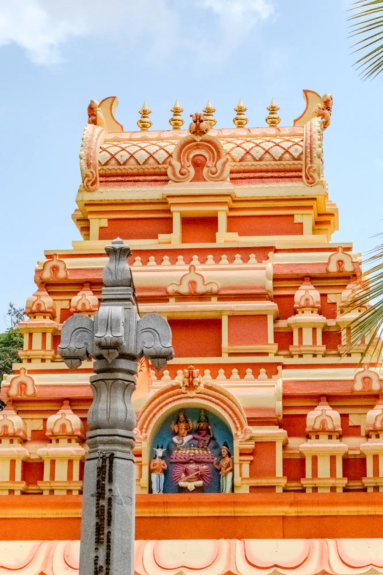 A multi-tiered Hindu temple tower rises in vibrant hues of saffron orange, apricot, and pale yellow against a light blue sky. Ornate horizontal moldings and stylized architectural motifs step upward toward a barrel-vaulted roof adorned with five golden finials along its crest. Near the base, a small arched niche houses sculpted polychrome figures set against a teal backdrop. In the foreground to the left, a tall, carved gray stone pillar with vertical inscriptions stands out in stark contrast to the saturated warm tones of the sanctuary.