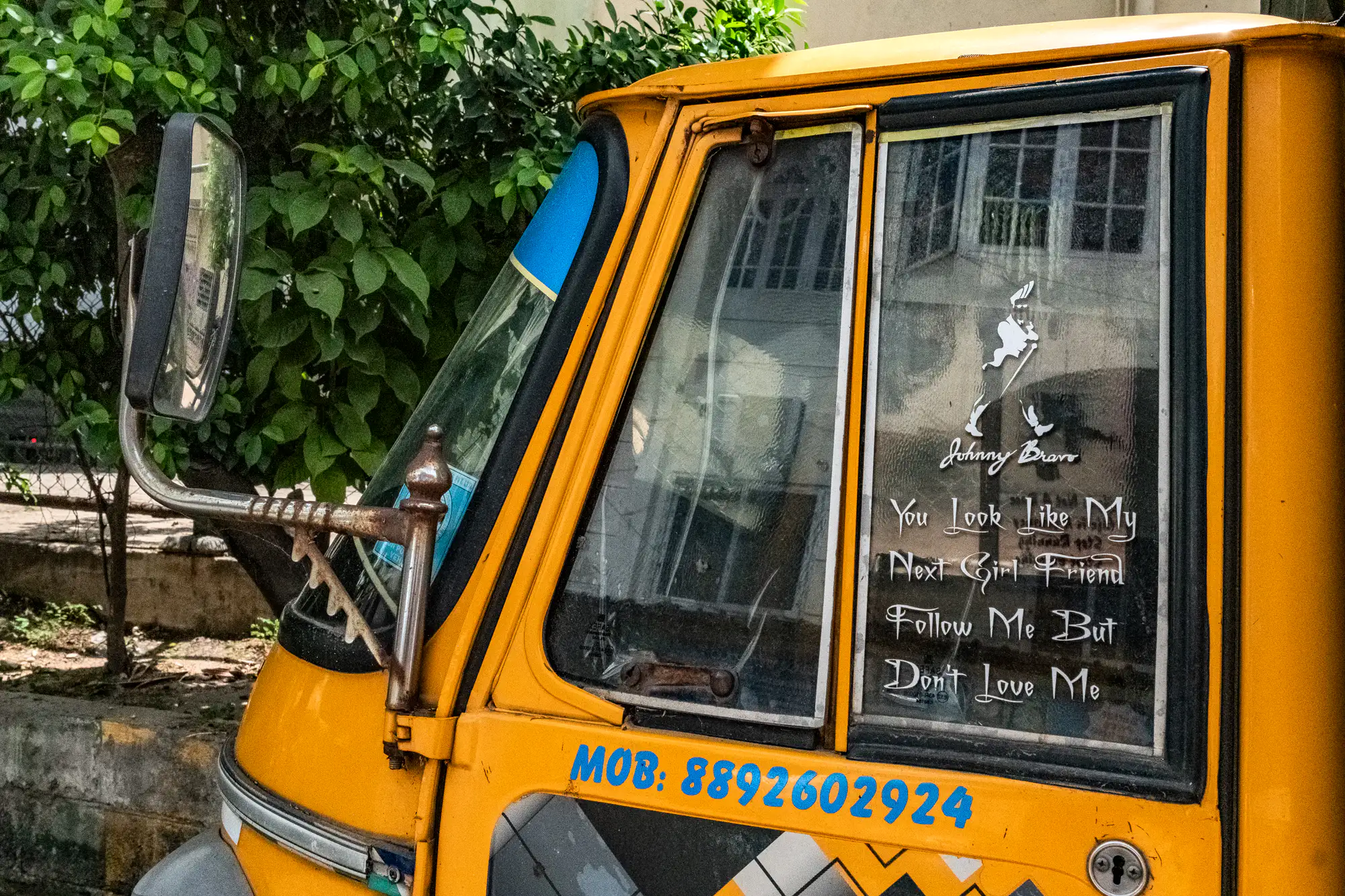 A bright yellow auto-rickshaw cabin fills the frame from an angled profile, detailed with handwritten-style window decals and weathered hardware. A rectangular side mirror extends forward from a rusted metal armature mounted beside the front windshield. Printed in white across the side window glass, playful text reads 'You Look Like My Next Girl Friend' and 'Follow Me But Don't Love Me,' crowned by a silhouette decal labeled 'Johnny Bravo.' Beneath the pane, a vibrant blue phone number is painted directly onto the yellow body, while leafy green foliage softens the outdoor background.