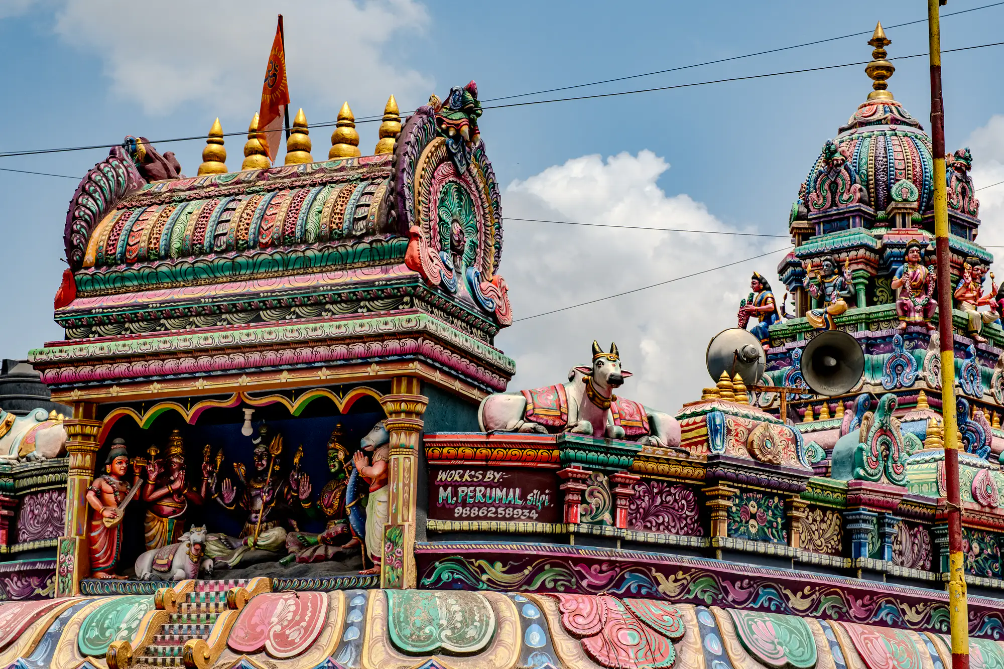 Intricately sculpted and vividly painted tiers of a Hindu temple rise against a bright blue sky dotted with soft white clouds. On the left, an open alcove shelters brightly colored divine figures beneath an ornate, barrel-vaulted roof capped with golden finials and an orange flag. A sculpted white bull rests on an embellished parapet beside mounted horn loudspeakers. To the right, a multi-tiered circular vimana tower, adorned with detailed deity figures and dense scrollwork in pastel and saturated tones, tapers upward toward a golden pinnacle.