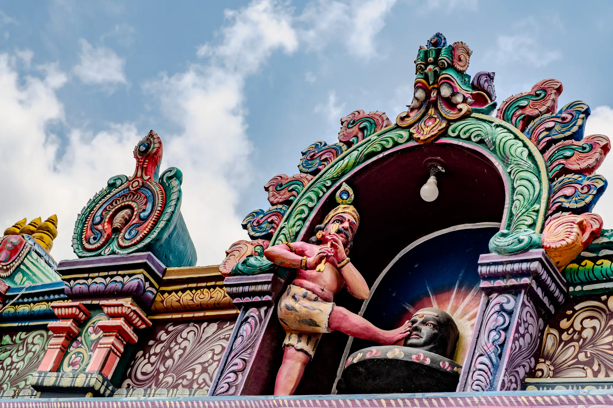 An ornate, vividly painted Hindu temple sculpture depicts a reddish-toned hunter figure standing within an arched alcove against a soft blue sky with scattered clouds. The figure, dressed in patterned shorts and wearing a feather headpiece, brings a golden arrow toward his eye while resting his extended foot against the sculpted face of a dark Shiva Lingam. Intricately carved ornamental flourishes in shades of green, coral, yellow, and violet frame the alcove, crowned with a demonic kirtimukha mask at its apex. An exposed modern light bulb hangs directly above the sculpted scene from the center of the dark interior niche.