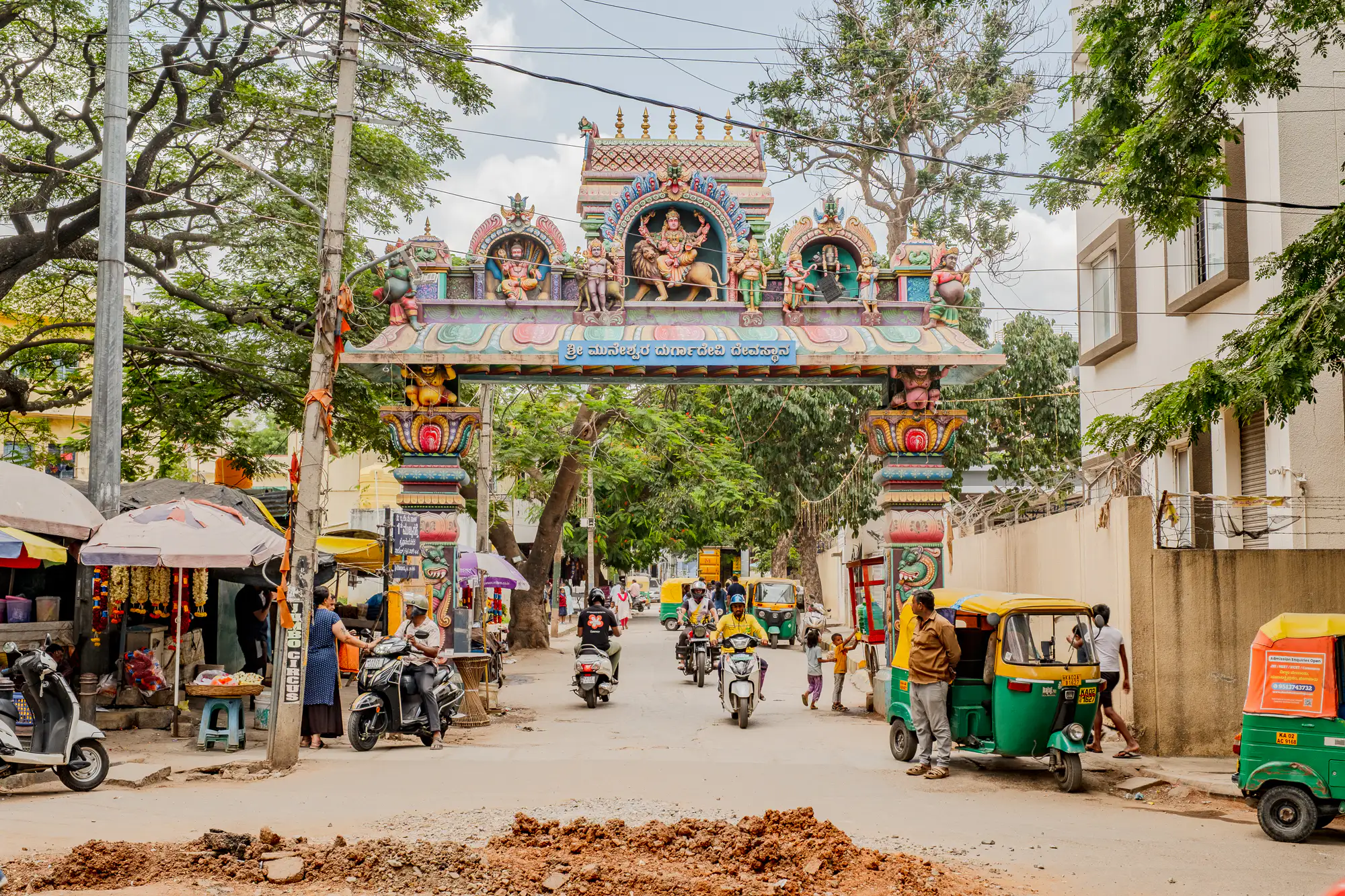 An ornate, multi-tiered Hindu temple archway painted in vivid polychrome shades spans across an active urban street. The decorative lintel showcases sculpted deities, anchored by a central niche depicting a multi-armed goddess astride a lion, supported by sculpted columns and figures below. Underneath the gate, auto-rickshaws, motorbikes, and pedestrians navigate the lane alongside shaded market stalls and crisscrossing utility wires. In the immediate foreground, a mound of excavated earth and rubble sits on the roadway under diffused daytime light.