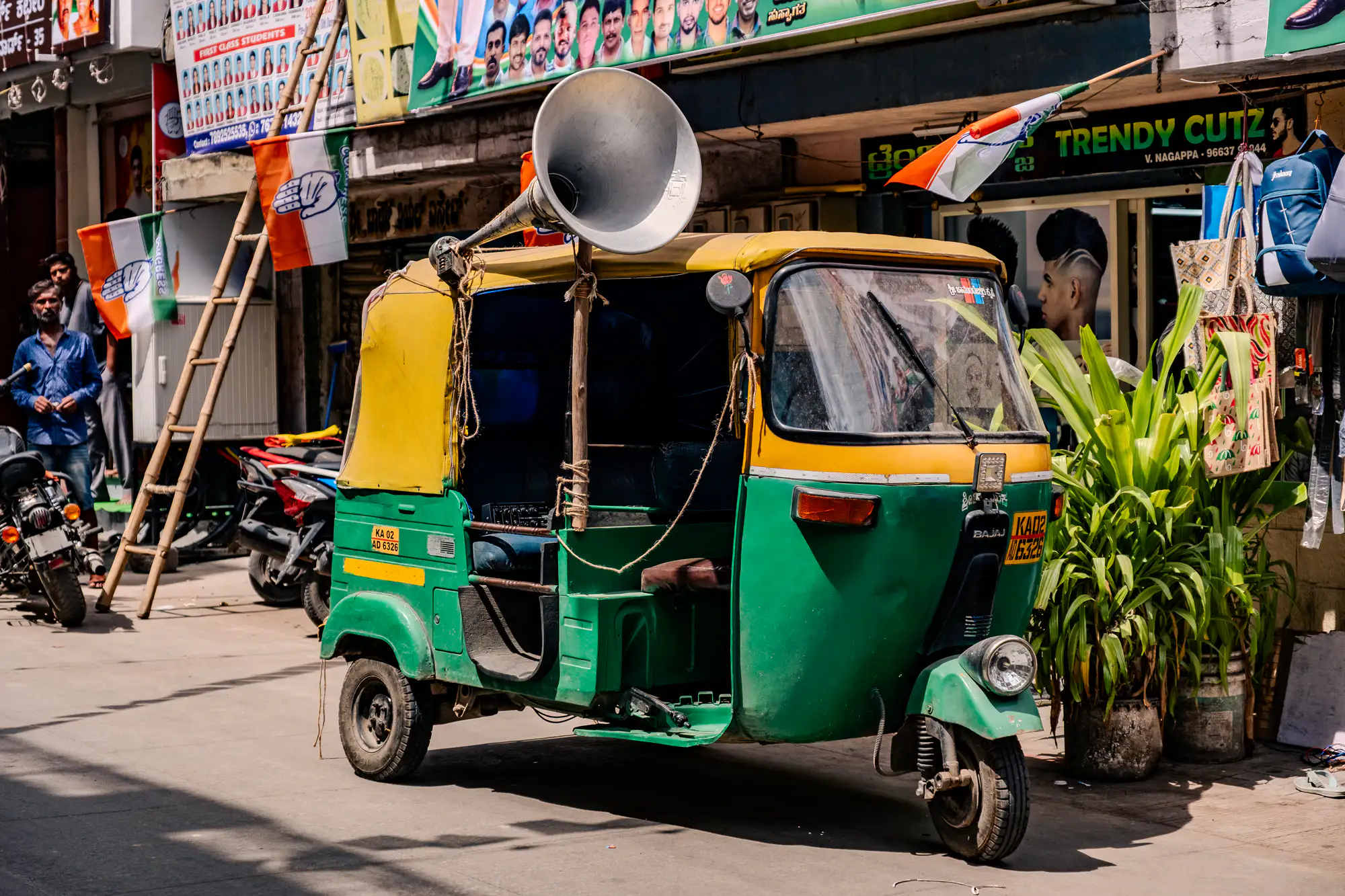 A green and yellow three-wheeled auto-rickshaw rests on a sunlit urban street in front of a shopfront. Fastened upright to the vehicle frame with ropes and a wooden post, a large silver loudspeaker horn faces forward above the cabin roof. In the background, tricolor flags showing an open palm symbol hang from a bamboo ladder alongside parked motorbikes and lush potted greenery. Crisp daylight casts defined shadows underneath the chassis, illuminating the textured street surface and worn exterior panels.