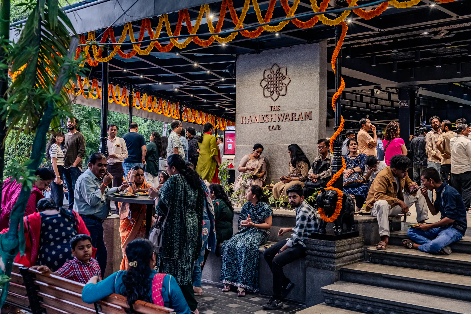 Patrons gather on stone steps and around high-top tables beneath the sheltered entryway of The Rameshwaram Cafe. Strands of bright orange and yellow marigold garlands drape along the ceiling beams and wind down a dark decorative pillar beside a small sculpted elephant. Groups of visitors dine from paper plates, converse, and wait along the tiered stone ledges under warm recessed lighting. Lush green foliage frames the open side of the bustling dining pavilion.