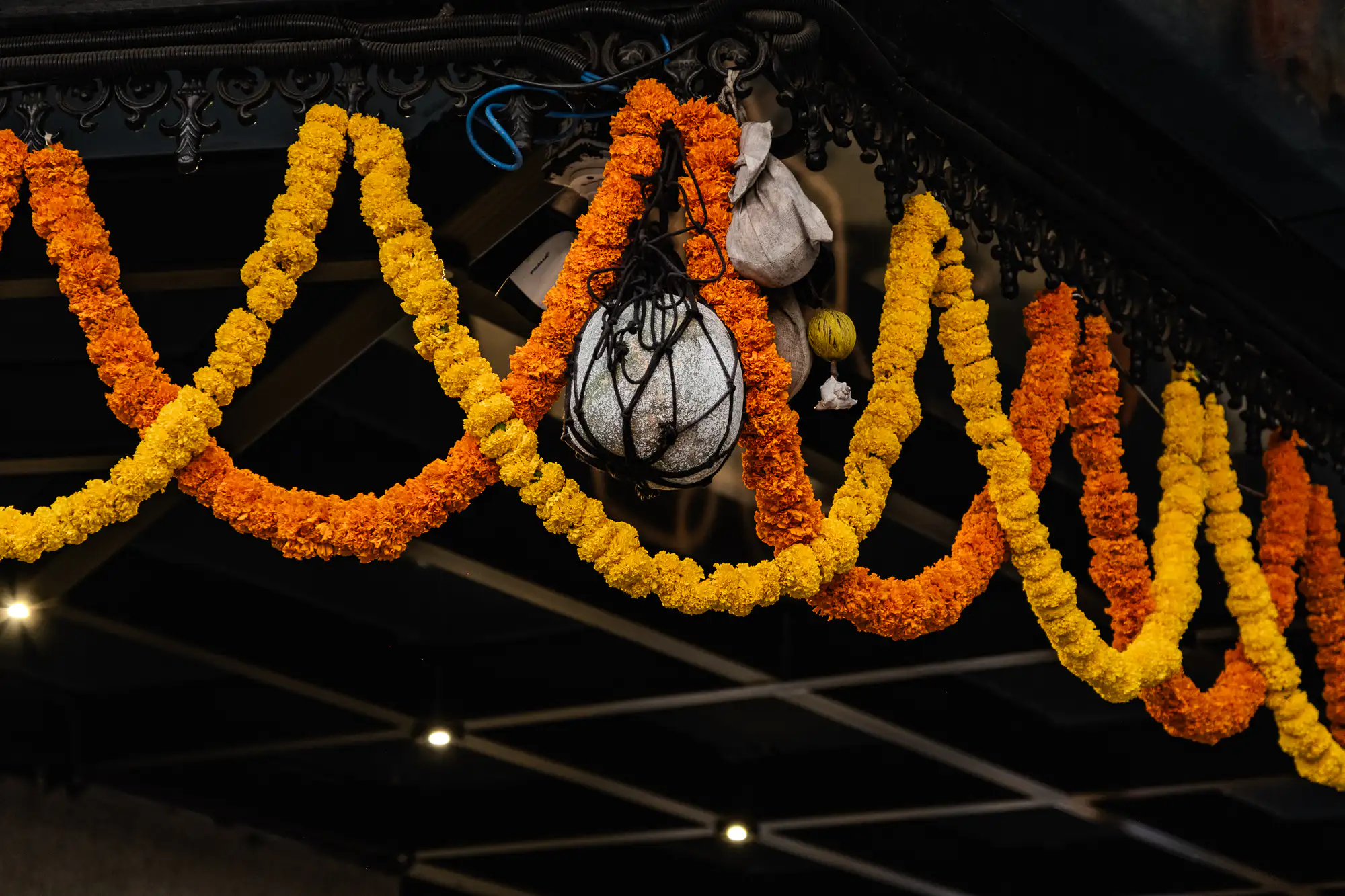 Vibrant strands of yellow and deep orange marigold garlands hang in sweeping swags from an ornate, dark architectural trim. Suspended at the center of the floral display is a pale ash gourd encased in black netting, accompanied by small cloth pouches and suspended charms. In the dimly lit background beneath the overhang, warm recessed ceiling lights glow against dark structural beams. A shallow depth of field keeps the dense floral textures and woven ropes in crisp focus against the shadowy interior.