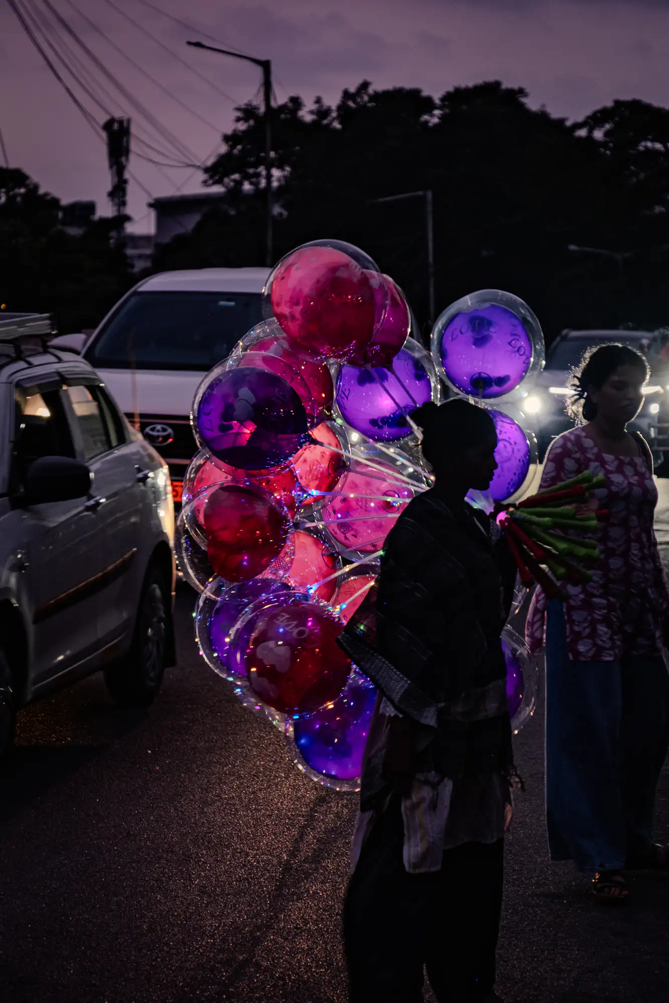 A street vendor silhouetted against evening twilight carries a large cluster of glowing transparent balloons filled with vibrant pink and purple spheres wrapped in tiny LED string lights. The vendor walks along an asphalt roadway flanked by passing motor vehicles whose headlights illuminate the street. Nearby, a pedestrian in a patterned pink tunic walks alongside as deep violet hues settle across the sky above silhouette tree canopies.