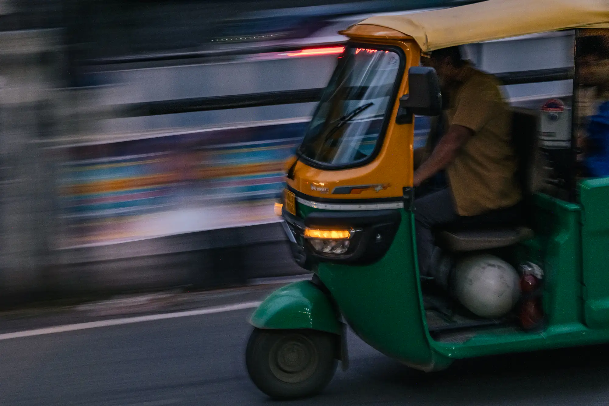 A green and yellow auto-rickshaw speeds along an asphalt road, captured in sharp focus through a dynamic panning technique. A warm amber indicator glows on its front grill, casting light forward as the driver steers from within the open-sided cabin. Due to the motion-tracking exposure, the surrounding urban backdrop melts into horizontal streaks of blurred color and cool evening tones.