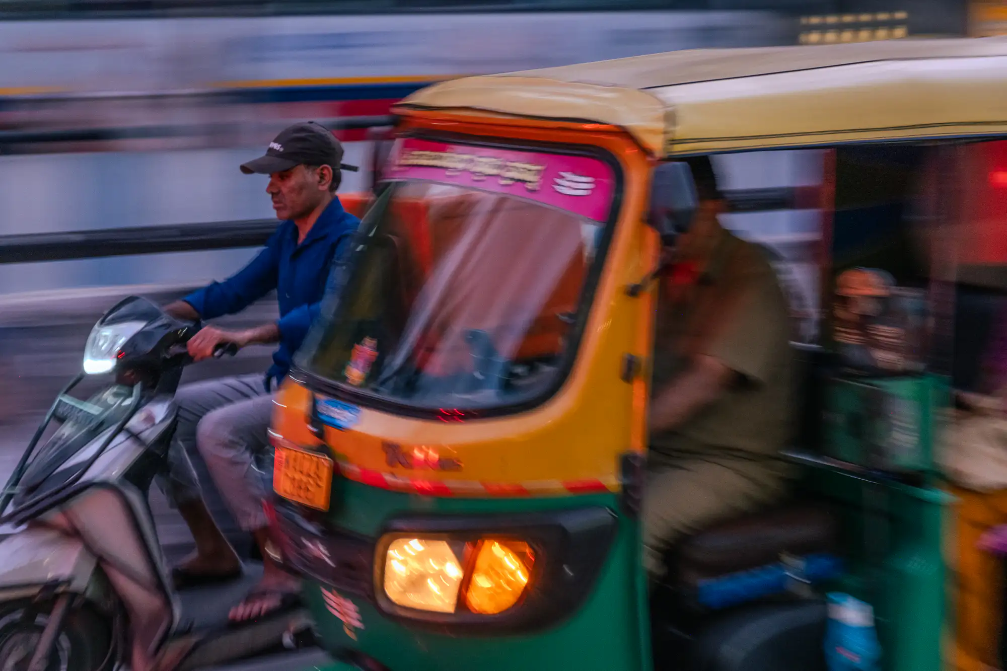 A vibrant yellow and green auto rickshaw speeds alongside a man steering a scooter along an urban roadway at dusk. Captured with a panning technique and slow shutter speed, horizontal streaks of blue, white, and violet blur across the background to emphasize forward momentum. The rickshaw's glowing amber headlight casts a warm beam against the cool evening light, while its driver sits partly visible inside the open-sided cabin. To the left, the scooter rider is depicted in sharp profile wearing a blue shirt and dark baseball cap, gazing intently ahead into traffic.