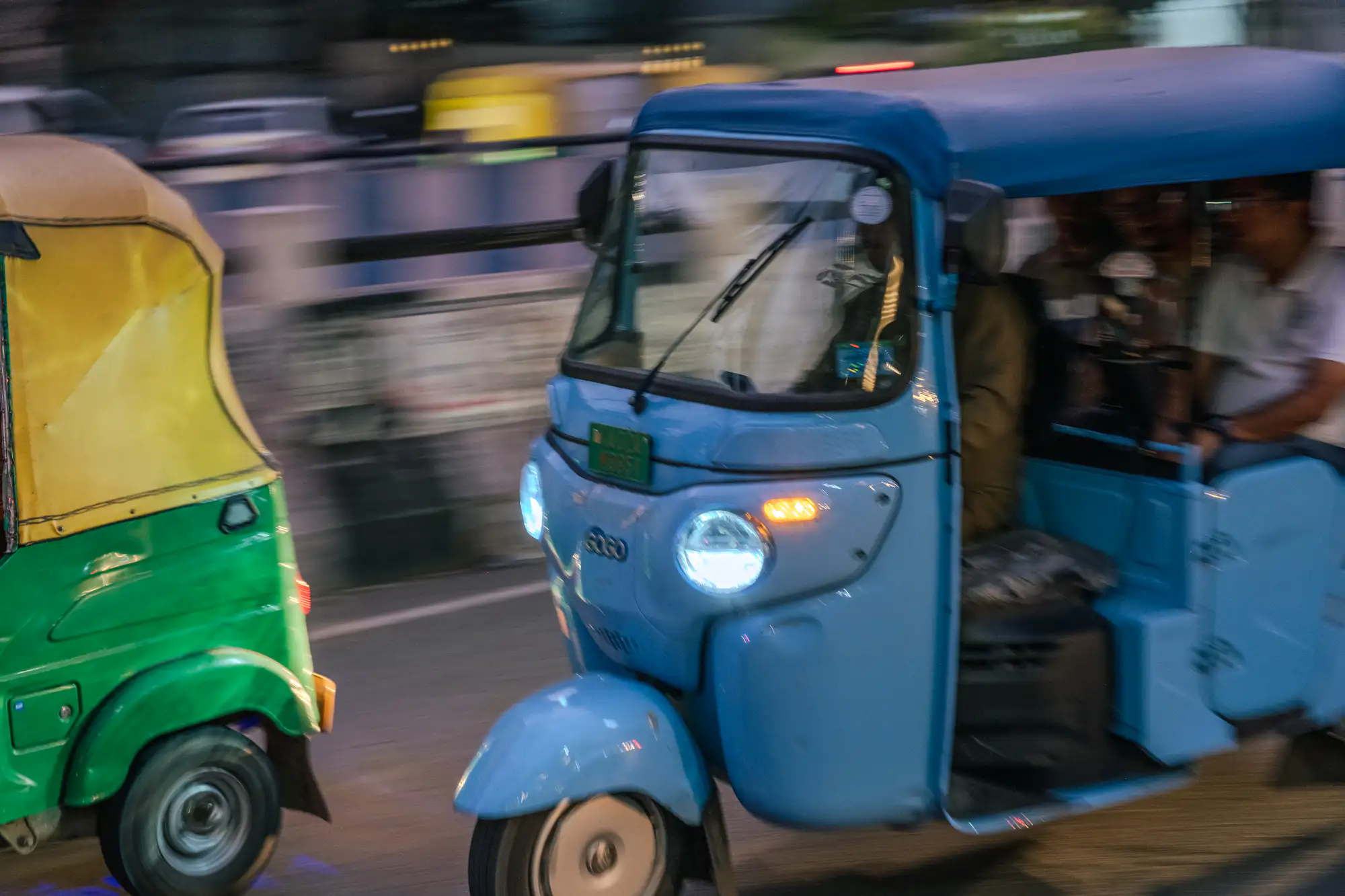 A bright blue three-wheeled auto-rickshaw speeds down an urban street, framed sharply against a heavily motion-blurred background of streaks and ambient city lights. The vehicle's front headlight emits a vibrant cool-toned glow next to an illuminated amber turn signal. Inside the open cabin, silhouettes of the driver and seated passengers remain partially visible, accompanied on the left by the rear half of a green and yellow auto-rickshaw traveling in parallel.