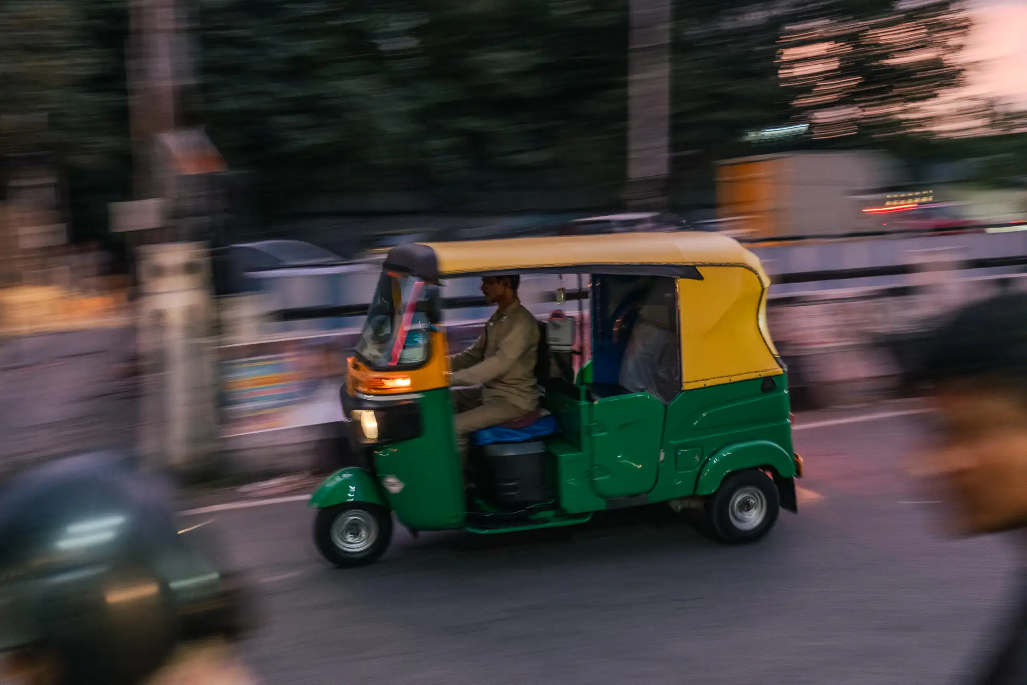 A green and yellow auto-rickshaw speeds across an asphalt street at dusk, rendered sharply against a heavily motion-blurred urban background. Inside the open-sided cabin, a driver in a tan uniform steers forward as the vehicle's amber front lamp casts a warm glow. Horizontal streaks of roadside barriers, distant foliage, and a soft pink twilight sky convey the swift panning motion. In the immediate foreground, the blurred dark shapes of passing commuters sweep past the lens.