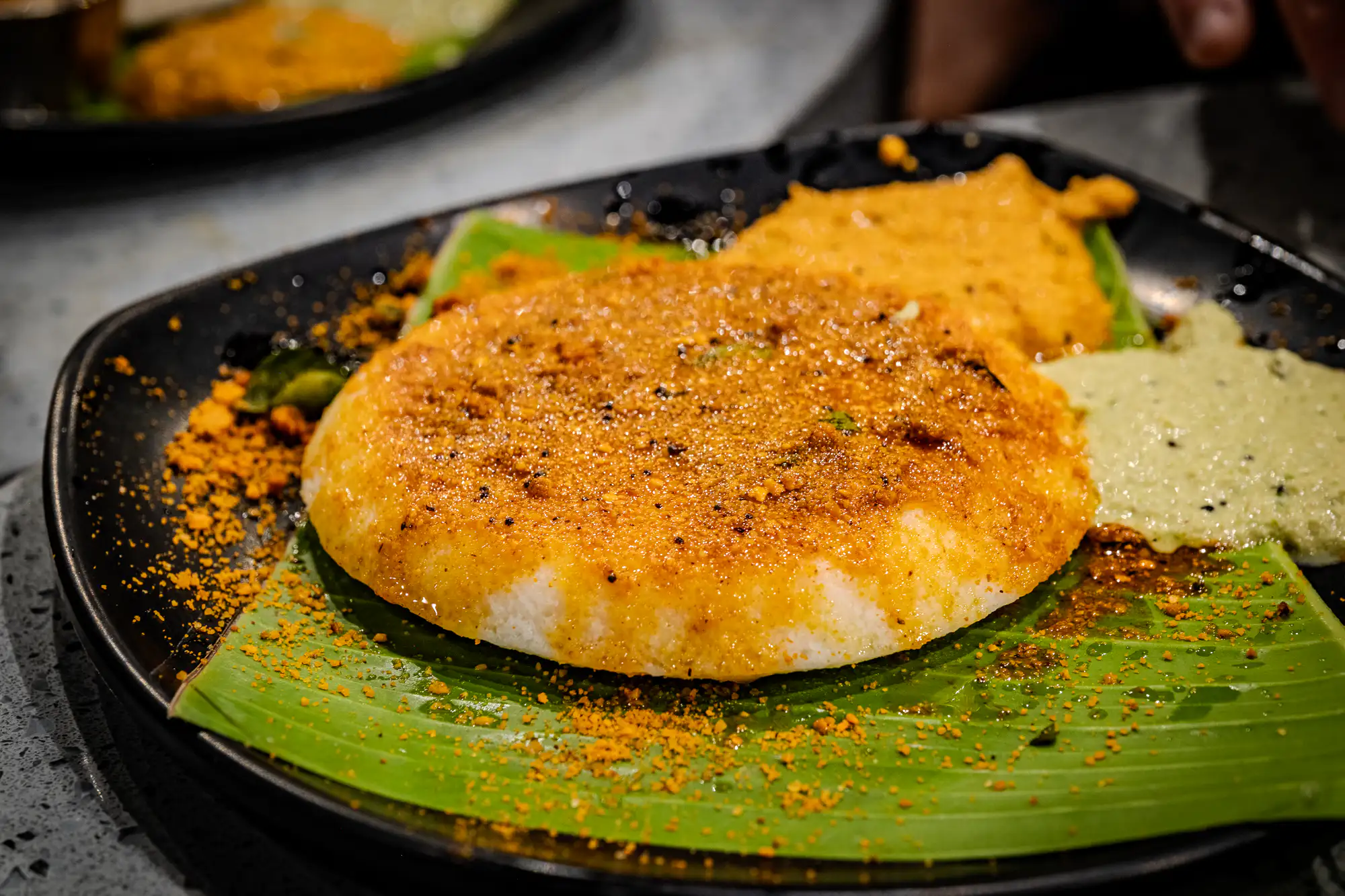 A thick, round steamed rice cake rests upon a vibrant green banana leaf lining a dark plate. The surface of the white cake is generously coated in a glistening layer of melted ghee and coarse, golden-orange spice powder that spills across the leaf. Alongside the cake sits a serving of pale green chutney speckled with mustard seeds and another dish partially visible in the upper frame. A shallow depth of field brings the textured, spice-dusted crust into sharp focus while softening the background dining setting.