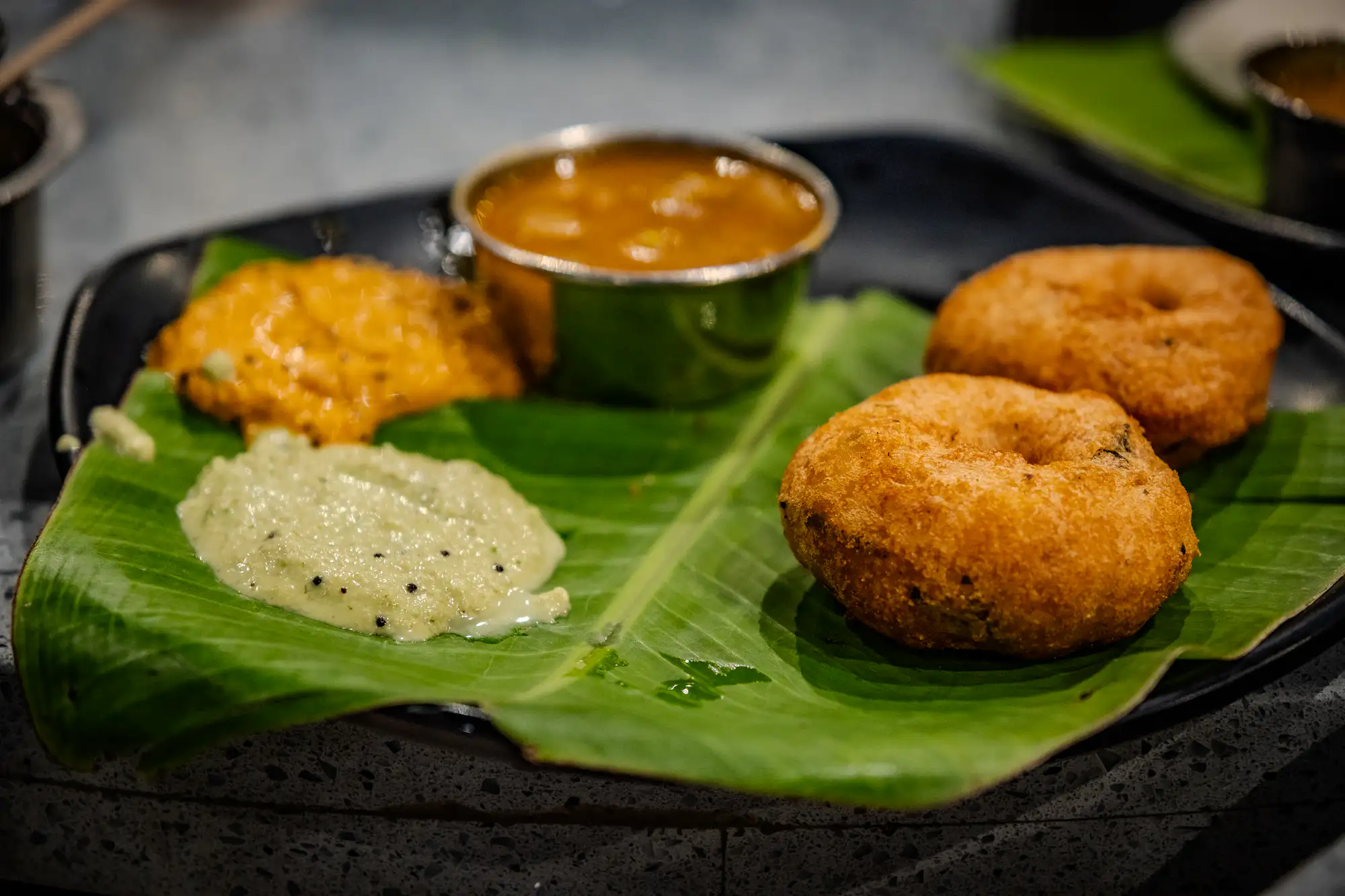 Two golden-brown, deep-fried medu vada rest on a vibrant green banana leaf cut to fit a dark plate. Beside them lie dollops of textured pale green coconut chutney speckled with mustard seeds and a bright orange-tinted chutney. A small, polished stainless steel bowl filled with amber-colored sambar sits at the center of the arrangement. A shallow depth of field keeps the foreground crisp while gently blurring the surrounding tabletop and adjacent dish.
