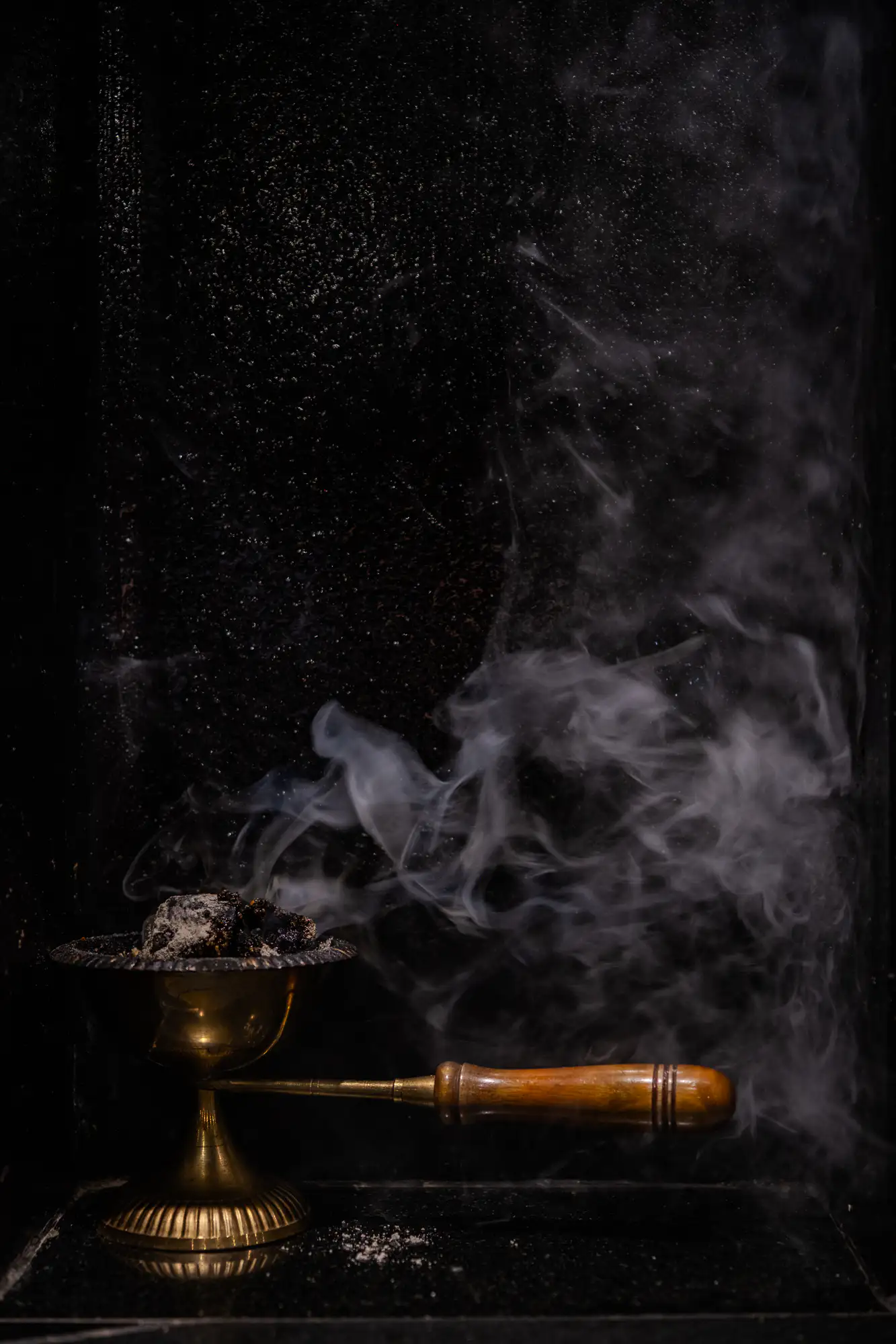 A brass incense burner with a carved wooden handle rests on a dark, reflective ledge. Inside its flared bowl, smoldering charcoal covered in pale gray ash releases dense, billowing swirls of white smoke. The semi-translucent plumes curl and disperse into the deep shadows of the textured black background. A scattering of fine ash dusts the dark surface near the ribbed pedestal base.