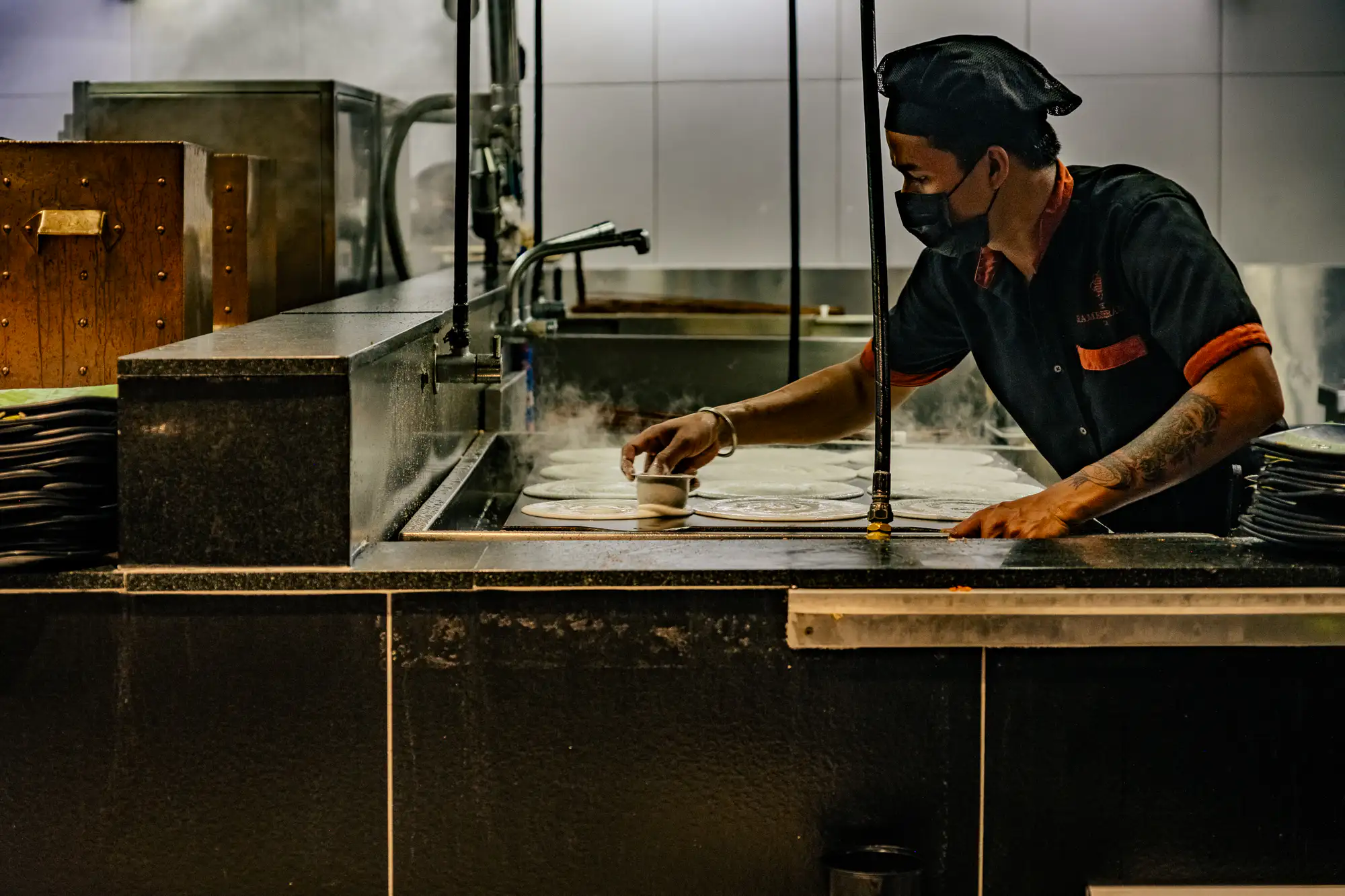 A cook wearing a black cap, a face mask, and a dark uniform with orange trim leans over a large flat-top griddle inside a commercial kitchen. Holding a small metal cup, his hand presses down to smooth thin, circular dosas across the heated surface as plumes of steam drift upward. Black granite countertops flank the cooking area, accompanied by neat stacks of plates and stainless steel fixtures against a tiled backdrop. Focused ambient lighting illuminates the precise motion of his craft while leaving the foreground in deep shadow.