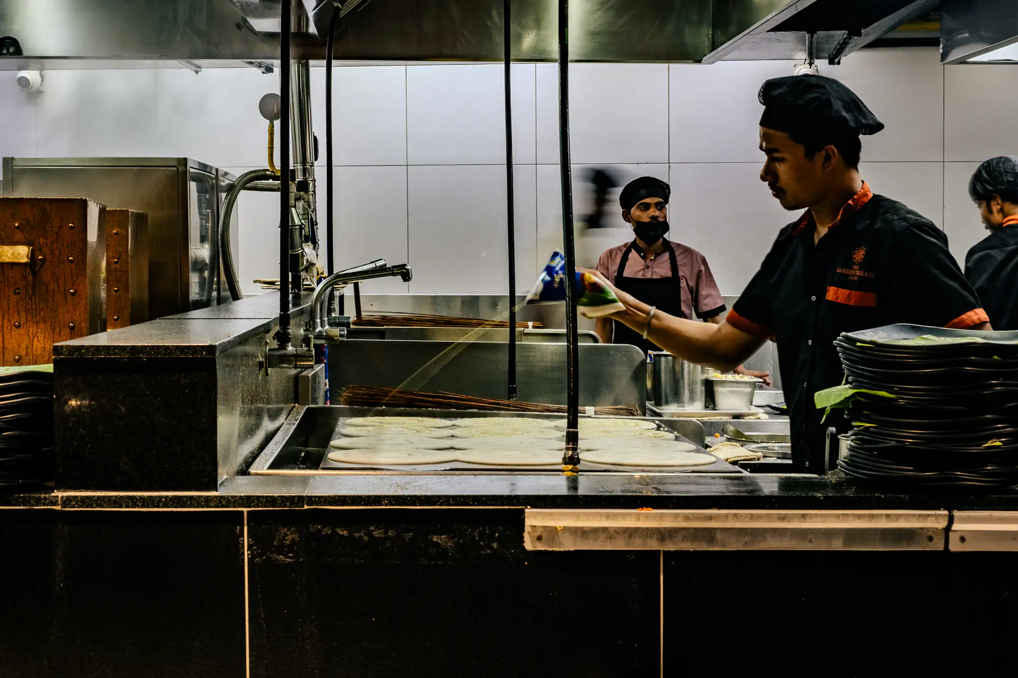 A cook wearing a black chef's cap and uniform pours a thin stream of liquid from a packet across rows of round dosas cooking on a broad flat griddle. In the foreground to the right, tall stacks of dark plates lined with green banana leaves rest on the stainless steel counter. Behind the cooking station, another staff member wearing a face mask observes the station while motion blur softens a passing figure in the background. Overhead illumination reflects off the dark granite partition, chrome faucets, and polished metallic exhaust hoods.