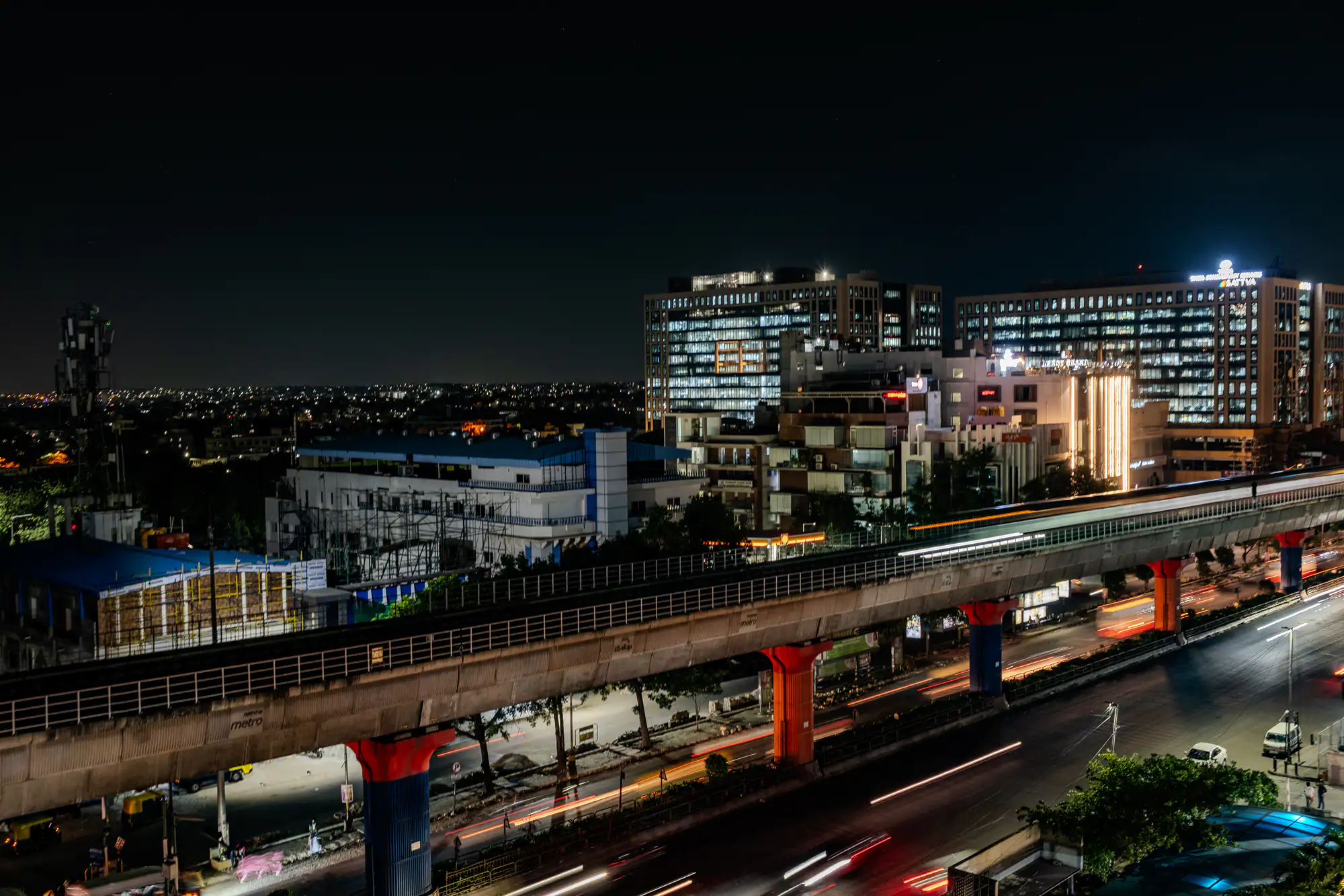 An elevated concrete transit viaduct spans diagonally across a bustling urban night scene, supported by large piers painted in vivid red and deep blue. A commuter train and moving road vehicles below appear as elongated streaks of white, gold, and red light produced by a long camera exposure. Modern, illuminated office towers and commercial complexes rise in the middle distance beneath a vast, pitch-black sky. Streetlights and building interiors cast a radiant, warm glow over the asphalt roadways and surrounding architecture.
