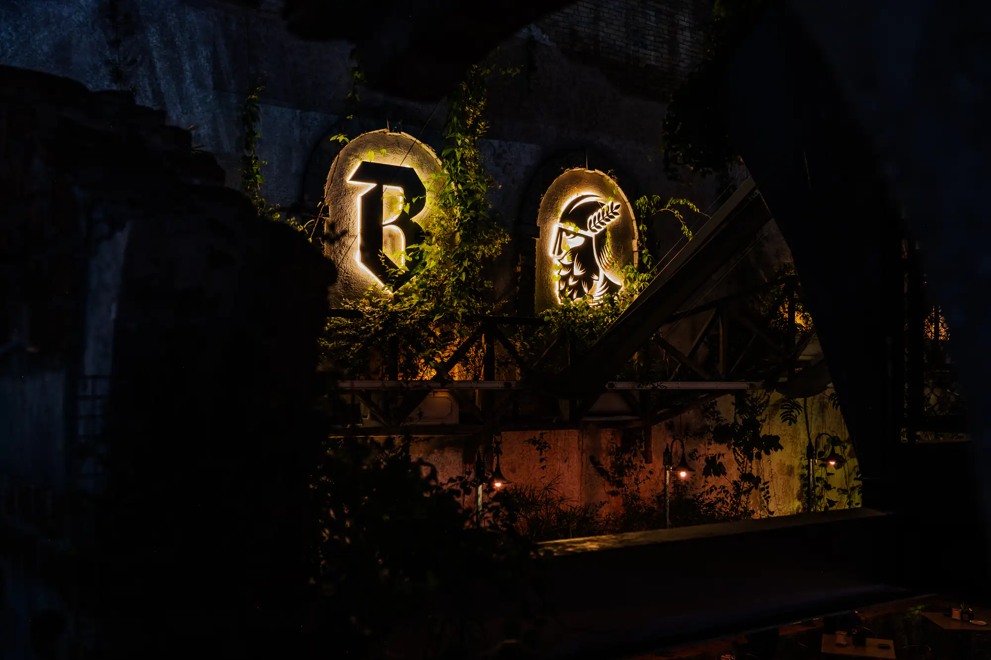 Two backlit emblems glow within arched masonry alcoves along a weathered, dark outdoor wall at night. On the left, an illuminated gothic letter 'B' stands out amidst climbing green ivy that partially drapes the stone facade. Beside it to the right, a second alcove displays a silhouetted profile of a helmeted figure adorned with a laurel wreath, radiant with warm white light. Beneath the arches, dark wooden scaffolding and dense foliage fall into heavy shadow, punctuated by small glowing filament fixtures.
