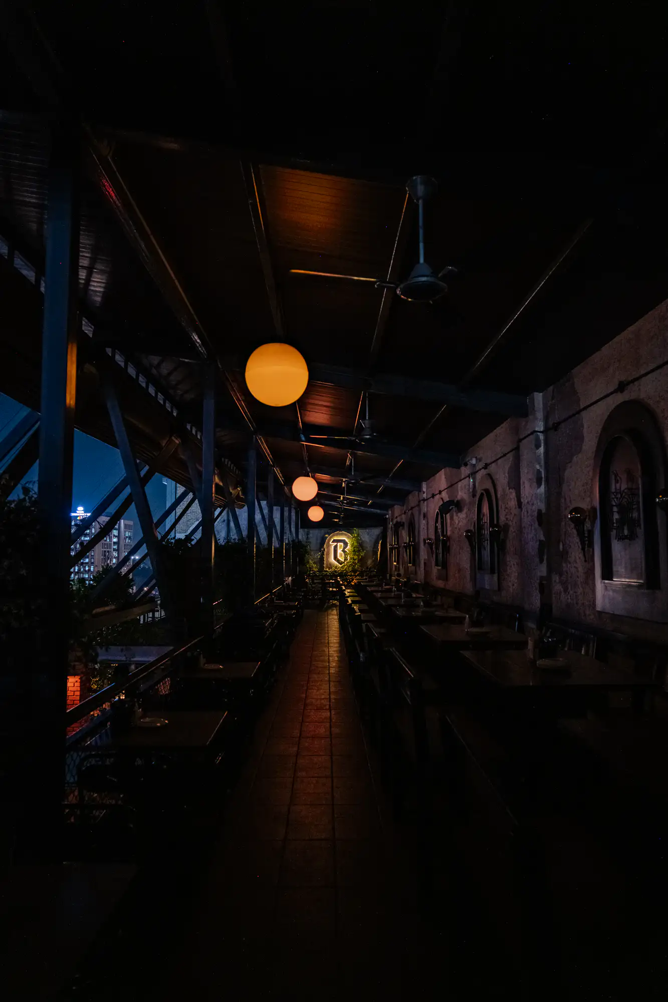A narrow, dimly lit covered terrace corridor extends into darkness under an angled open-beam ceiling. Luminous, amber spherical pendant lamps hang in a straight line overhead, illuminating the central tiled walkway and leading toward a backlit shield emblem at the far end. Rows of empty dining tables line the shadowed pathway, framed by textured walls with arched niches on the right and dark structural steel trusses on the left overlooking distant city lights.