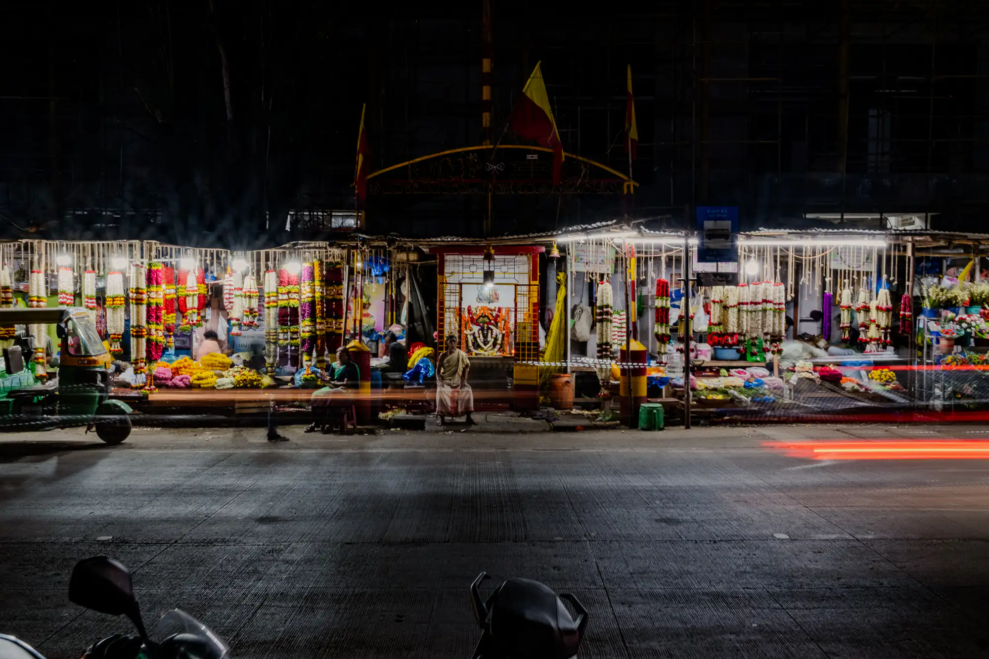 A roadside flower market and central illuminated shrine glow brightly against the darkness of a nocturnal city street. Long exposure photography renders horizontal streaks of red and white light from passing traffic across the lower half of the storefronts. Dense curtains of vibrant yellow, red, and white flower garlands hang suspended under fluorescent canopy lighting where vendors and seated residents rest on the sidewalk. In the immediate foreground, the dark handlebars and mirrors of a parked scooter frame the textured asphalt roadway.