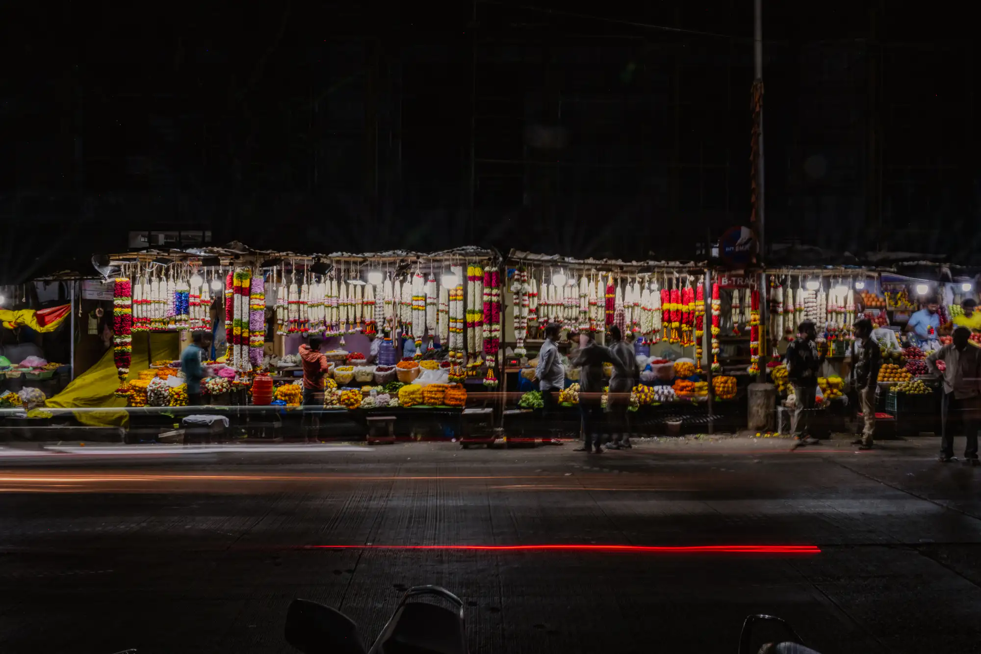 An outdoor flower market stall glows intensely beneath a line of hanging bulbs against an otherwise black night sky. Densely arranged floral garlands in saturated shades of yellow, white, and deep red hang in vertical curtains above piles of fresh blossoms displayed across open counters. Several figures linger along the edge of the stall, their outlines softened into subtle motion blur from a one-second exposure. In the foreground, horizontal streaks of brilliant white and crimson light trace across the dark, paved street from passing traffic.