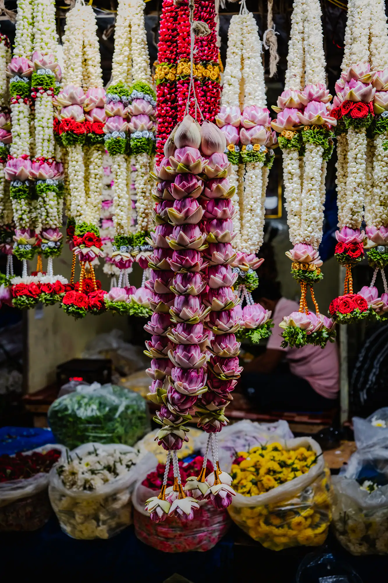 Hanging floral garlands fill the frame of a market stall, centered around a dense, vertical column of folded pink lotus buds. Flanking the central lotus garland are long strands of tightly woven white jasmine interspersed with pink petals, vibrant red roses, and beaded tassels. Below the suspended displays, large open sacks overflow with loose yellow marigolds, white blossoms, and red floral remnants. A shallow depth of field leaves the foreground garlands crisply defined against the shadowed stall interior where a seated figure works.