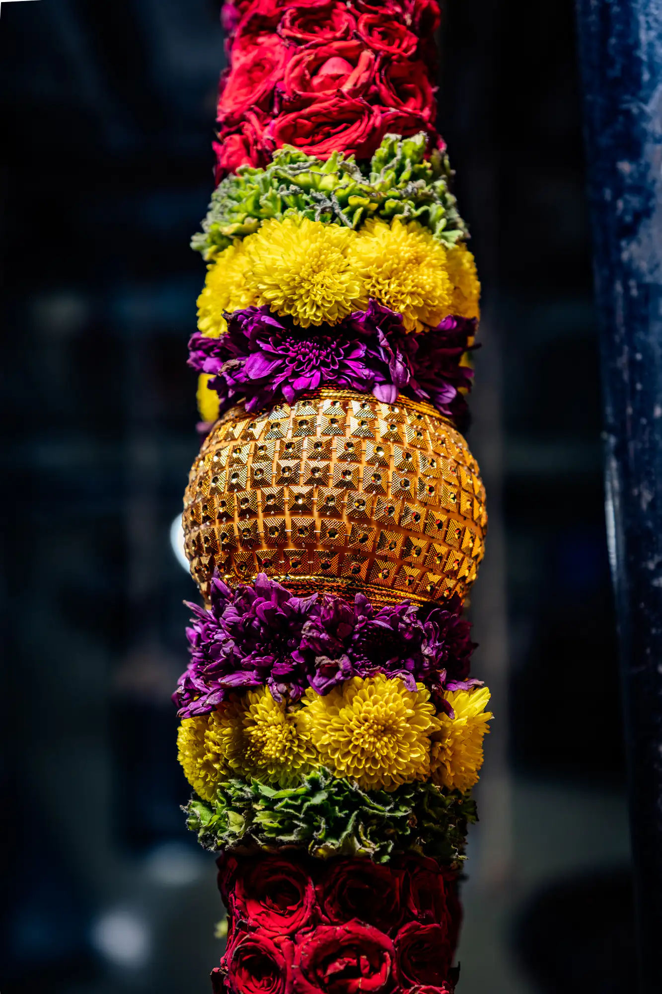A vertical floral garland column features a central spherical golden ornament surrounded by symmetrical tiers of brightly colored fresh blossoms. The textured metallic sphere is embellished with raised square studs and bordered immediately on both sides by dense rings of deep violet petals. Radiating outward from the violet layers are bands of vibrant yellow pompon chrysanthemums, fringed with ruffled green foliage and capped by clusters of deep red roses. A shallow depth of field isolates the ornate, handcrafted column against a dark, softly blurred background.