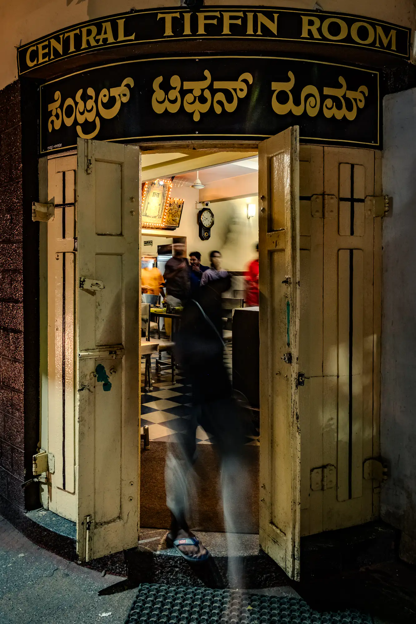 Pale yellow wooden double doors stand open beneath a black arched sign bearing the words 'CENTRAL TIFFIN ROOM' in English and Kannada script. A long exposure causes a figure passing through the threshold to appear as a dark, translucent streak of motion blur, with only a blue sandal briefly grounded on the entryway. The illuminated interior beyond displays a blue-and-white checkerboard floor, dining tables, patrons, and an illuminated shrine on the wall. Warm light from inside spills out onto the dark pavement and a textured rubber entrance mat below.
