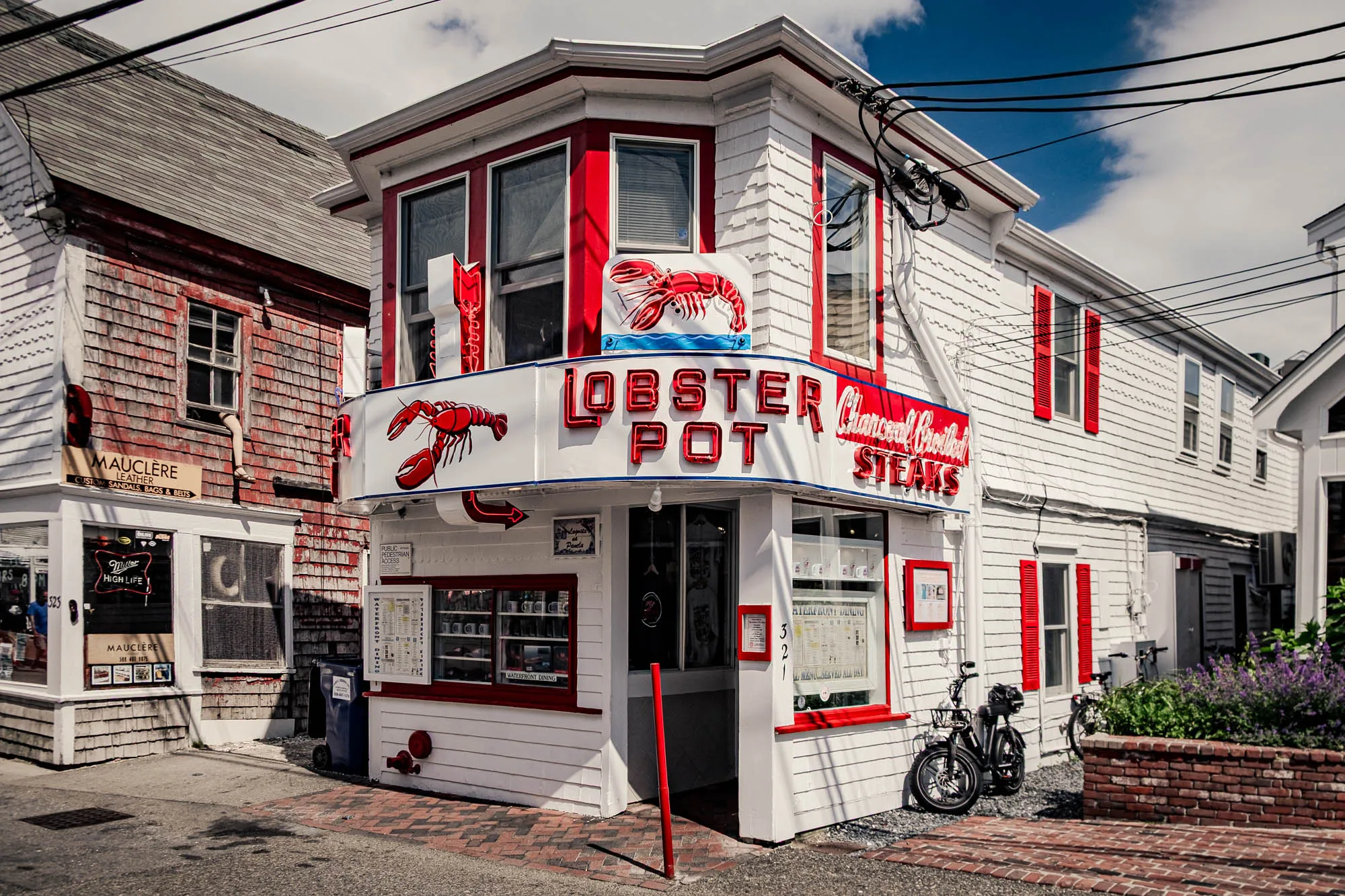 The image shows the exterior of a restaurant called "Lobster Pot". The building is primarily white with red accents, including the window trim, shutters, and neon signage. The restaurant sign is a prominent feature, displaying the name "LOBSTER POT" in large, red neon letters. Above the name, a neon lobster illustration stands out. Below the sign and around a corner entrance is a display case featuring menus and various items, framed in red. To the left of the restaurant, there is another building made with what appears to be old red cedar shingles which houses "Mauclere Leather". A small Miller High Life advertisement can also be seen in the window. A section of the brick sidewalk and street is visible in the foreground. Power lines crisscross the sky above the buildings, which is partly cloudy.