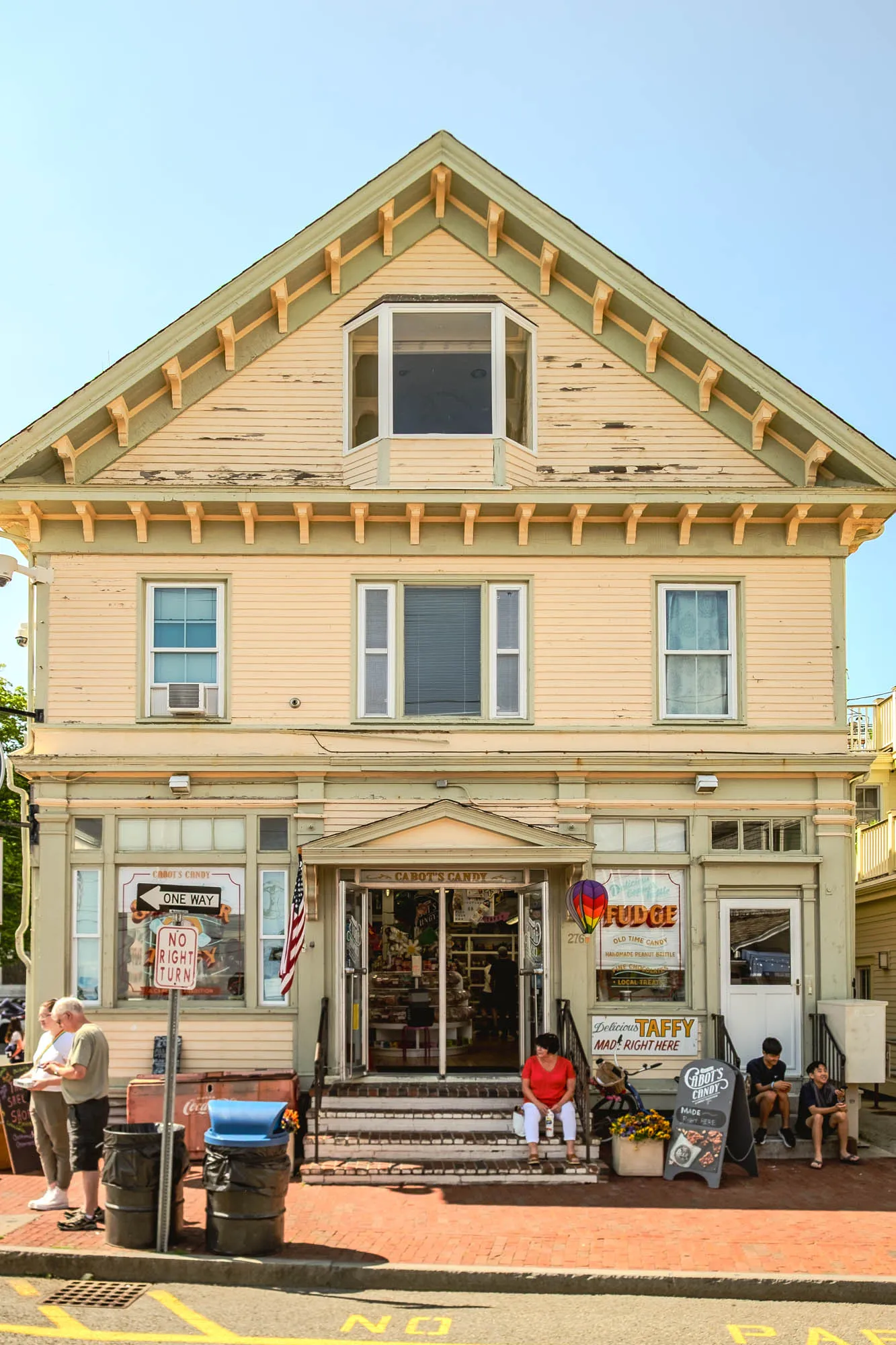 The image shows a two-story, clapboard-sided building under a clear, bright blue sky. The building's paint, a faded yellow with light green trim, shows signs of age, with some areas peeling. A large triangular pediment tops the building, featuring a centrally placed bay window on the second story and decorative brackets along the eaves. Two rectangular windows flank the bay window, one on each side, and another two windows are visible on the lower level. The windows have light-colored frames and some have drawn blinds. The building is a storefront, with a prominent sign reading 'Cabot's Candy'. An American flag hangs next to the entrance. In front of the store, several people are visible. To the left, an older couple stands near some trash cans and a sign indicating 'One Way' and 'No Right Turn'. On the steps leading into the store, a woman sits, while on the right, two young boys are sitting on the steps. A chalkboard sign advertising 'Cabot's Candy, Made Right Here' is also visible near the boys. The street is paved with red bricks and yellow traffic lines are on the pavement.