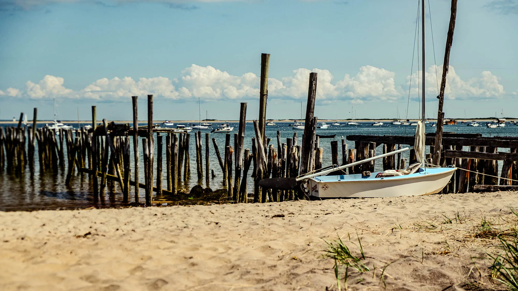The photograph shows a serene coastal scene on a bright day. In the foreground, a sandy beach slopes upward, dotted with sparse tufts of green grass. To the right, a small, light-colored sailboat rests on the sand, its mast extending upwards into the sky. Behind the boat, an old wooden pier stretches out into the water, its weathered pilings standing like sentinels in the sea. The pilings are dark with age and encrusted with marine growth. In the distance, the calm blue water is dotted with numerous sailboats and small motorboats, creating a lively seascape. Across the water, a low, sandy shoreline is visible, adding depth to the scene. The sky above is a clear, vibrant blue, adorned with puffy white clouds that add a sense of peace and tranquility to the overall composition. The scene evokes a sense of calm, a nostalgic view of a seaside town. The light is strong and warm, suggesting a summer day.